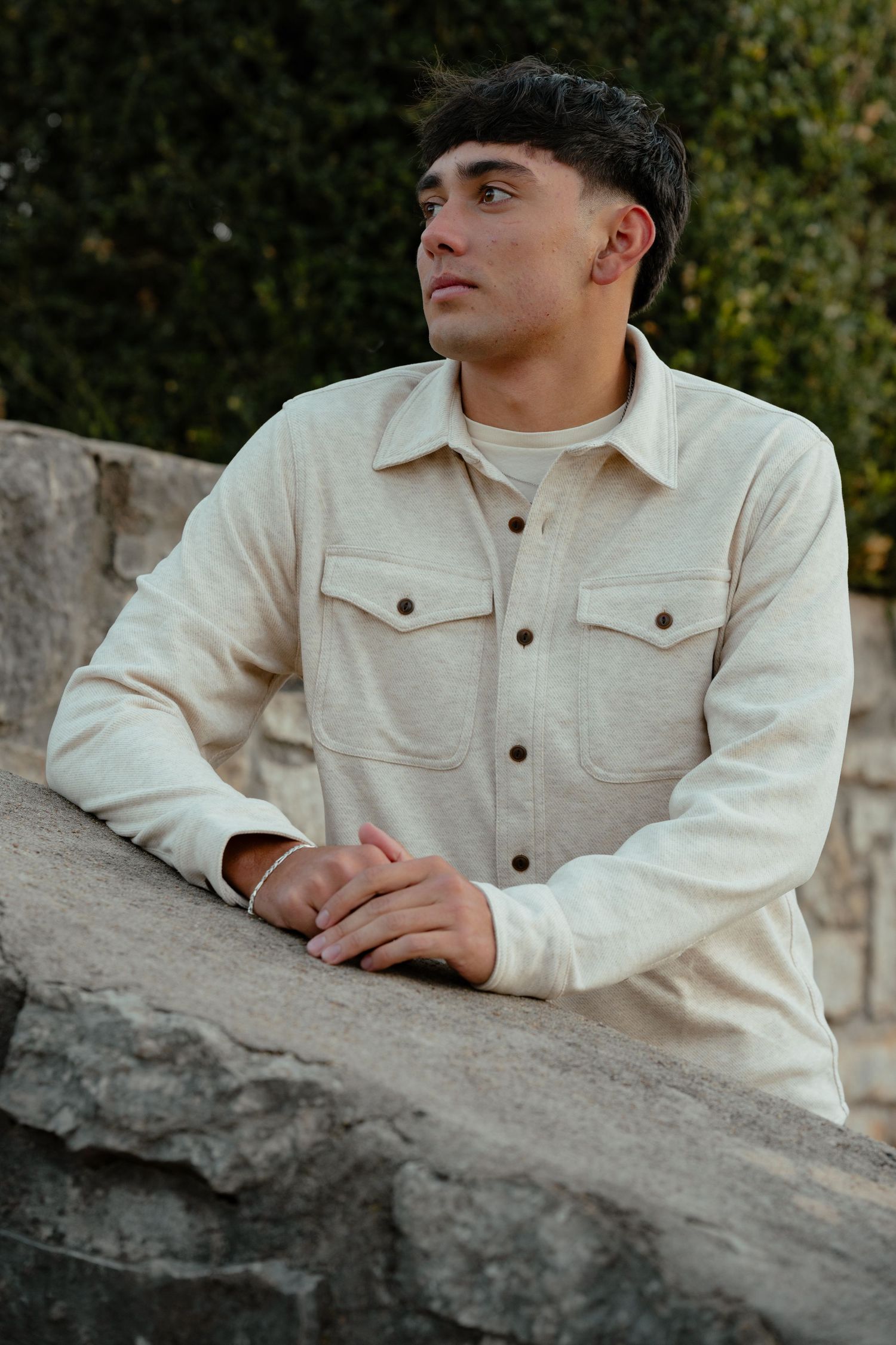 Side profile portrait against a stone wall wearing a cream-colored jacket in natural lighting.