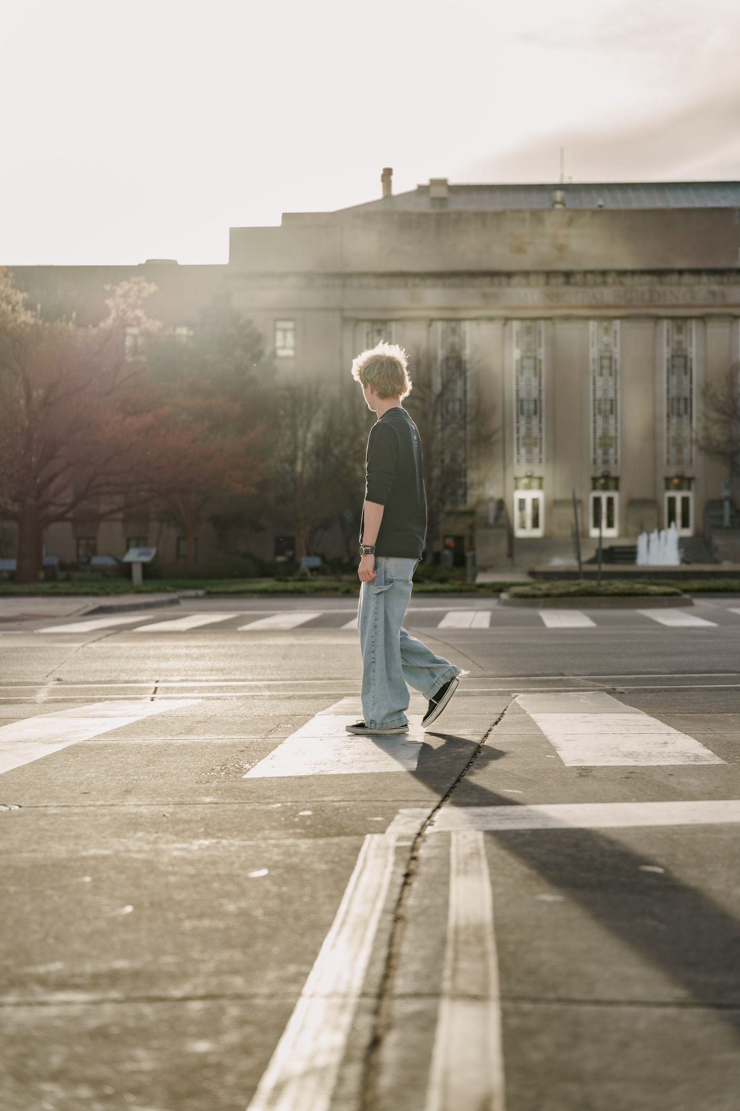Young person with blonde hair crossing a sunny urban crosswalk in wide-leg jeans near a classic stone building.