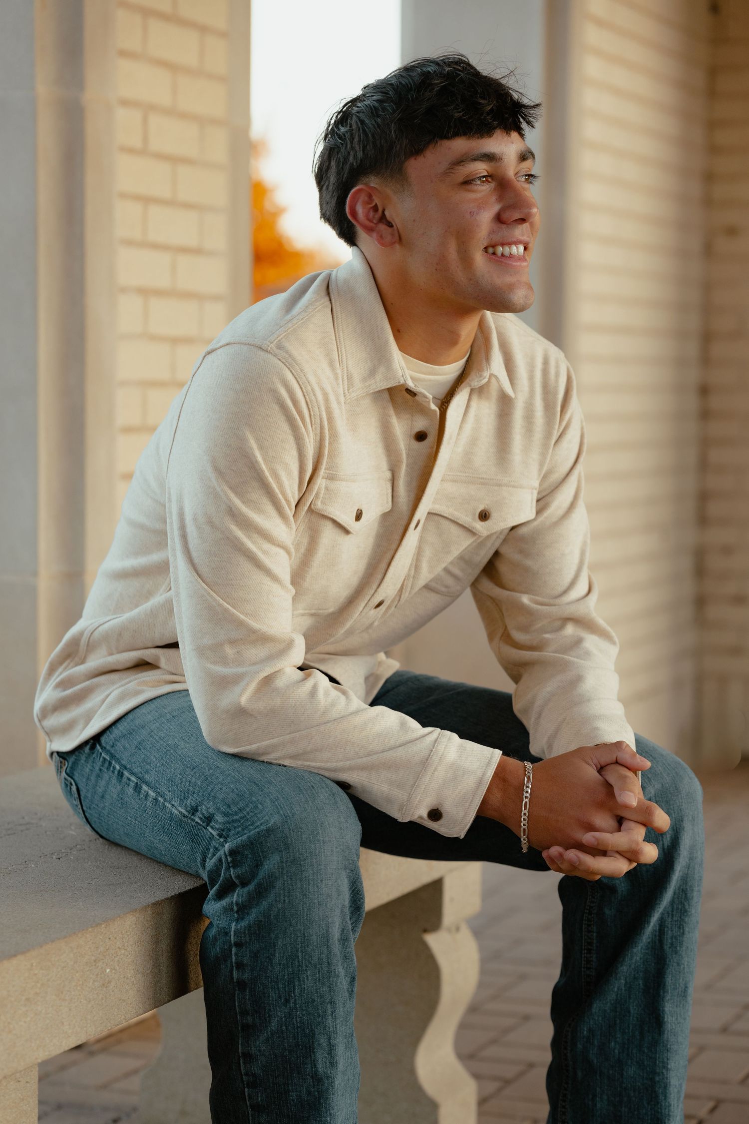 A casual portrait showing someone seated wearing a cream jacket and jeans in an architectural setting.