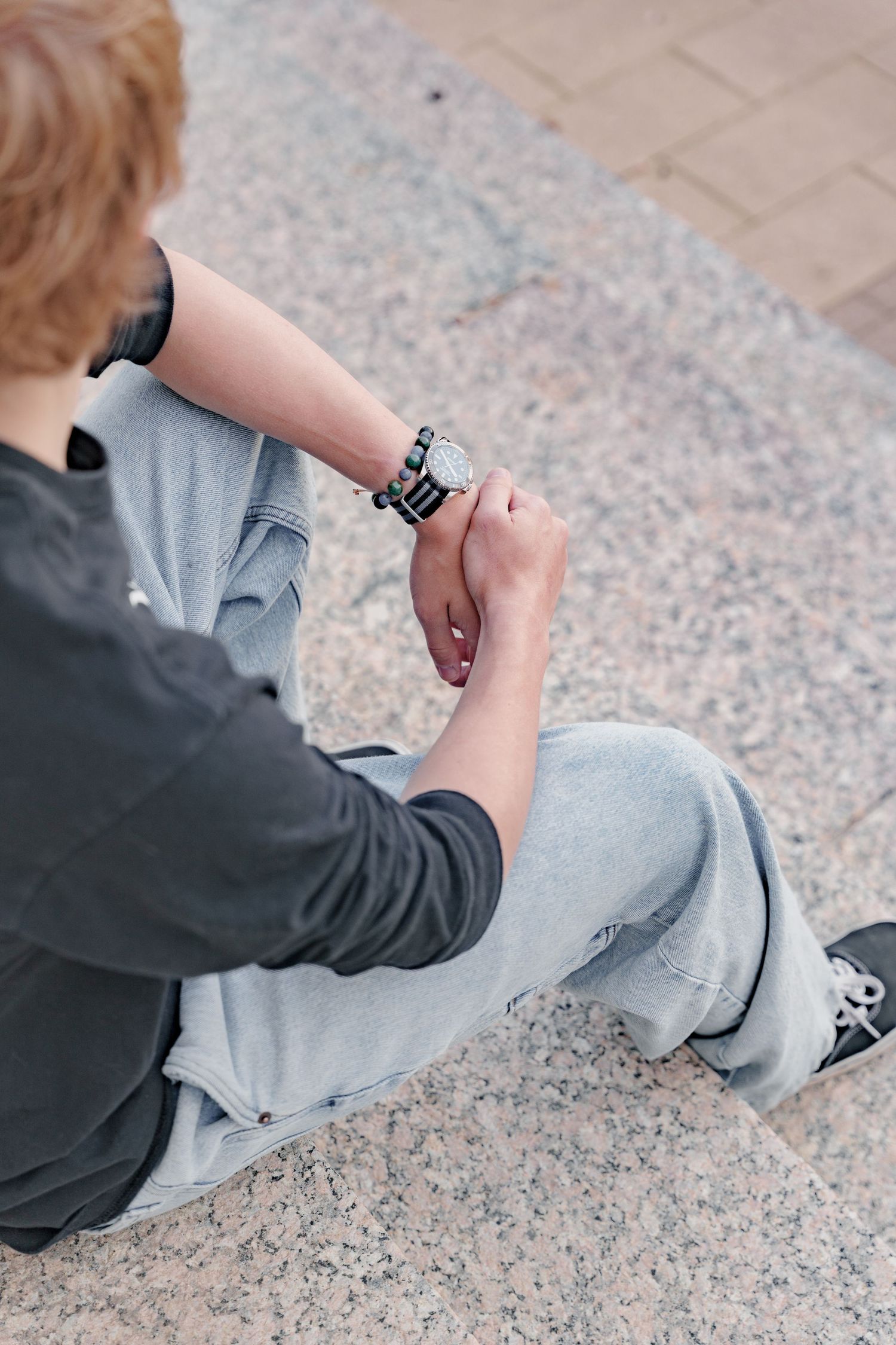 Young person sitting on granite steps checking their wristwatch, wearing gray jeans and dark jacket.