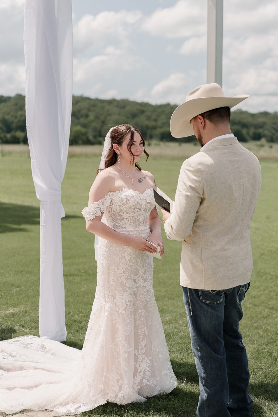 Bride and groom exchange vows outdoors at a rustic wedding ceremony, groom wearing a cowboy hat and linen blazer.
