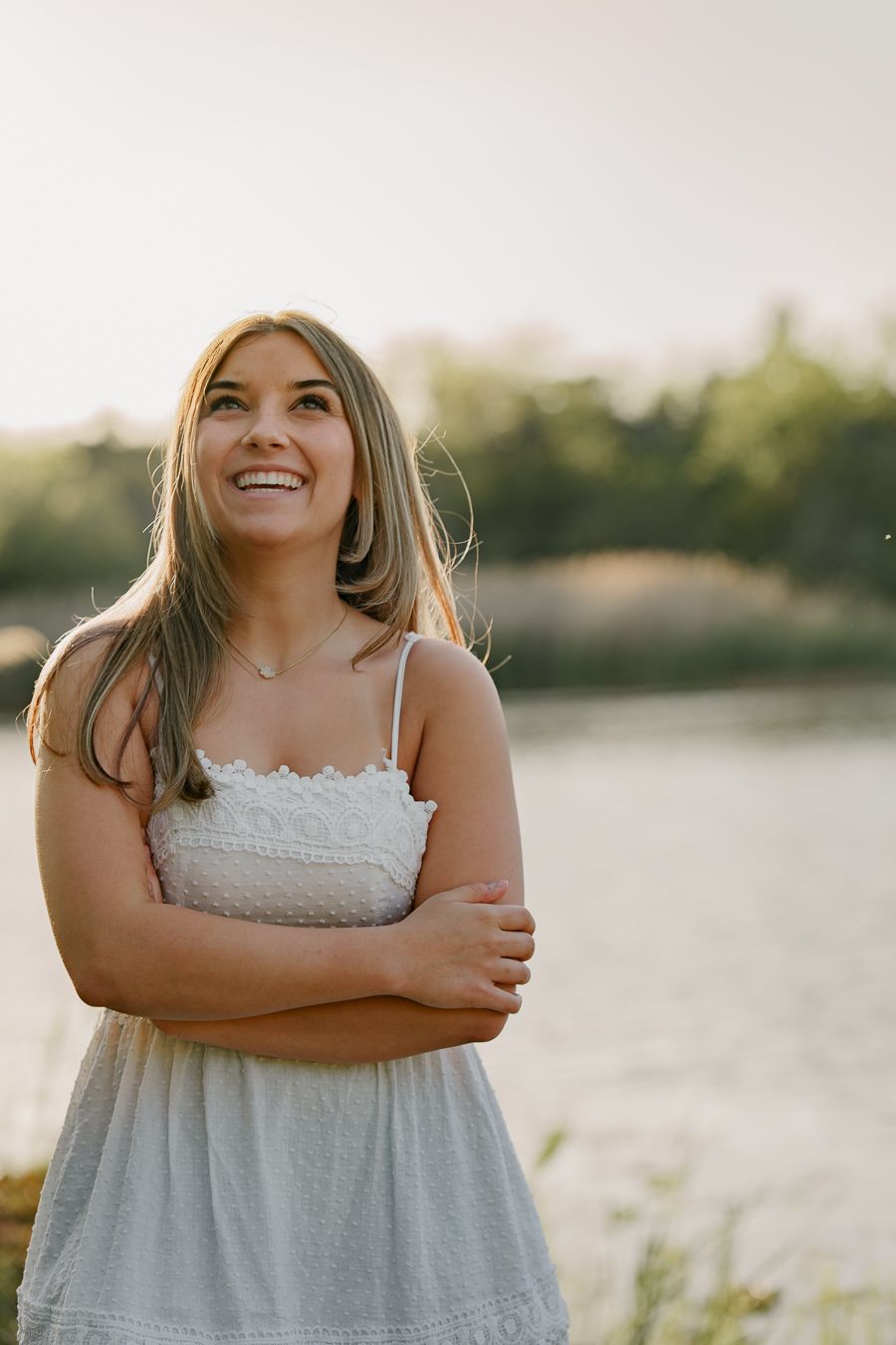 Young woman in white sundress smiling outdoors near a lake at golden hour with trees in background.
