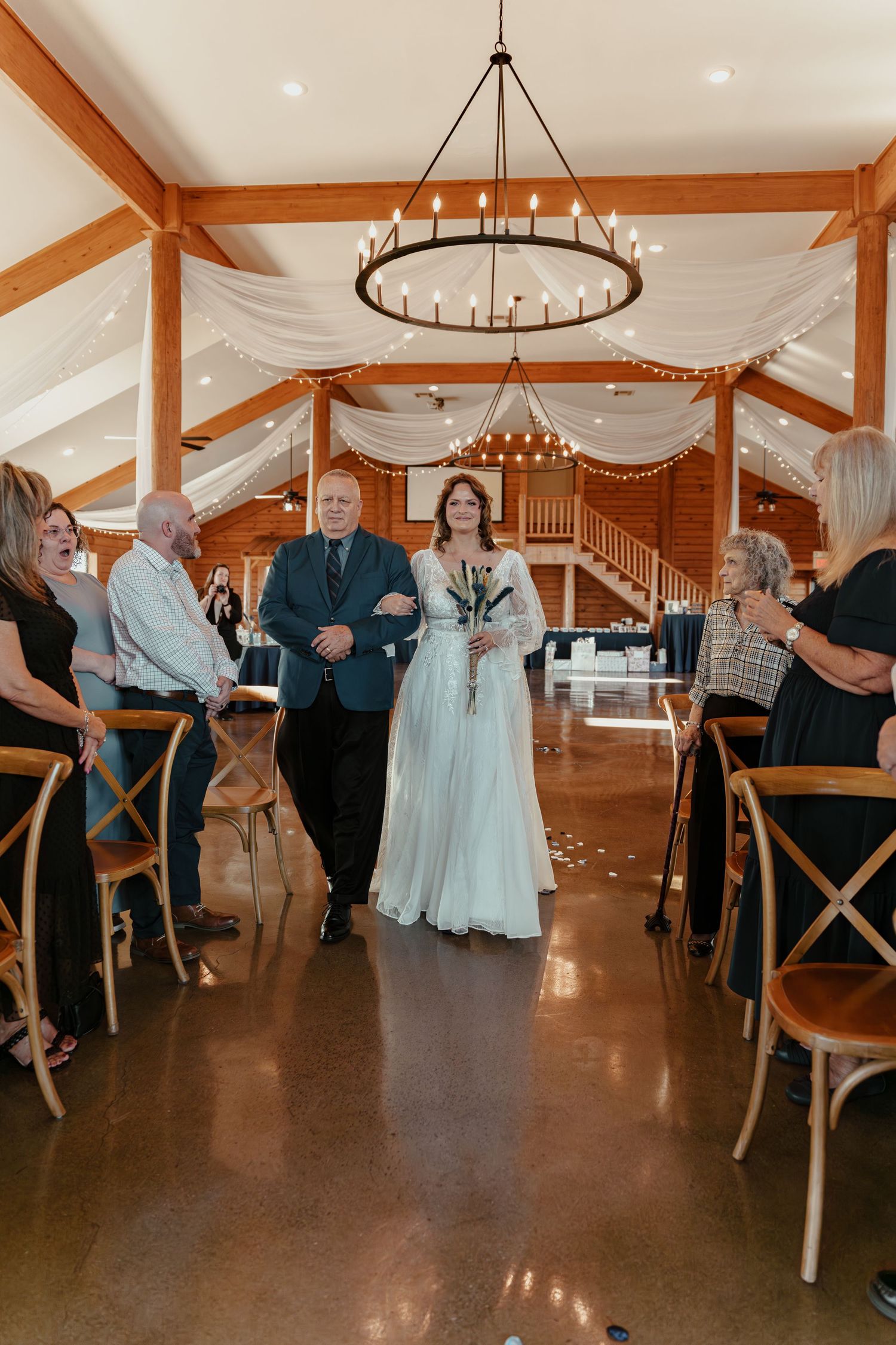 Bride walking down the aisle with escort in elegant barn wedding venue with chandelier and draped fabric decor.