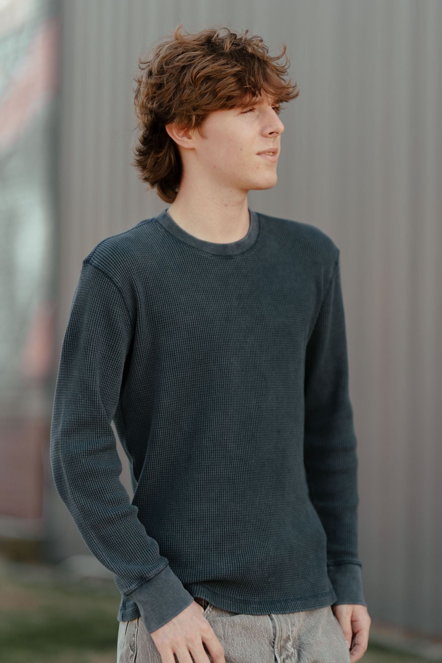 Young man with curly hair wearing a dark gray knit sweater standing outdoors against a metal wall.