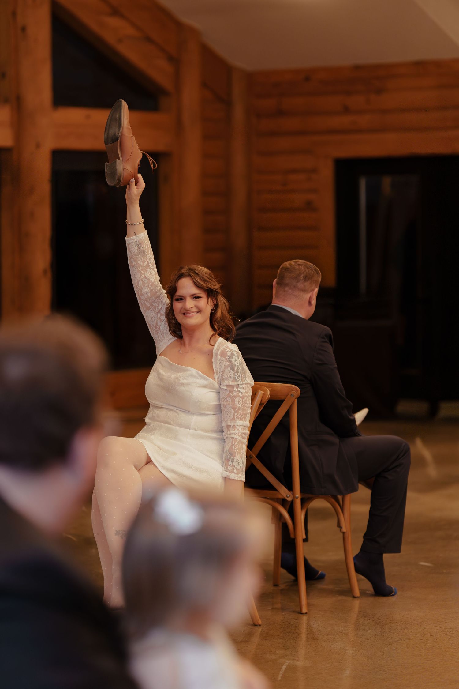 Bride in white lace dress raises groom's shoe during fun wedding game at rustic wood-paneled venue.