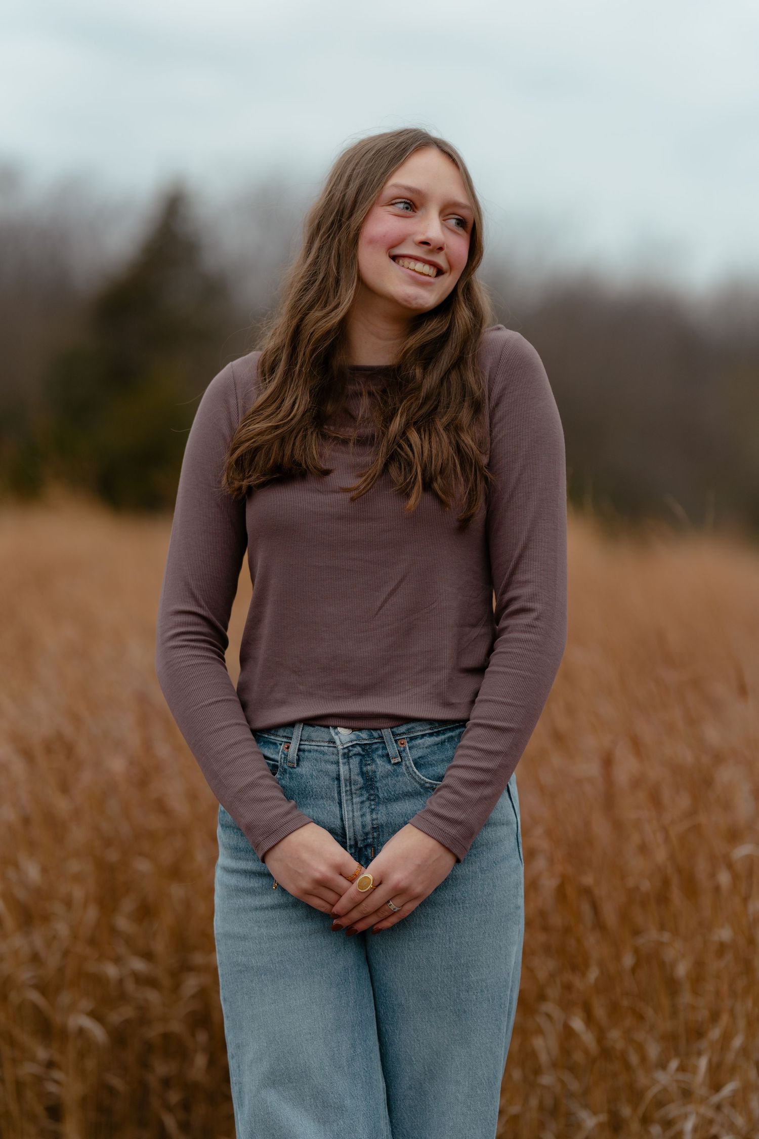 Young woman in brown long-sleeve top and jeans smiling in a golden field with trees in the background.