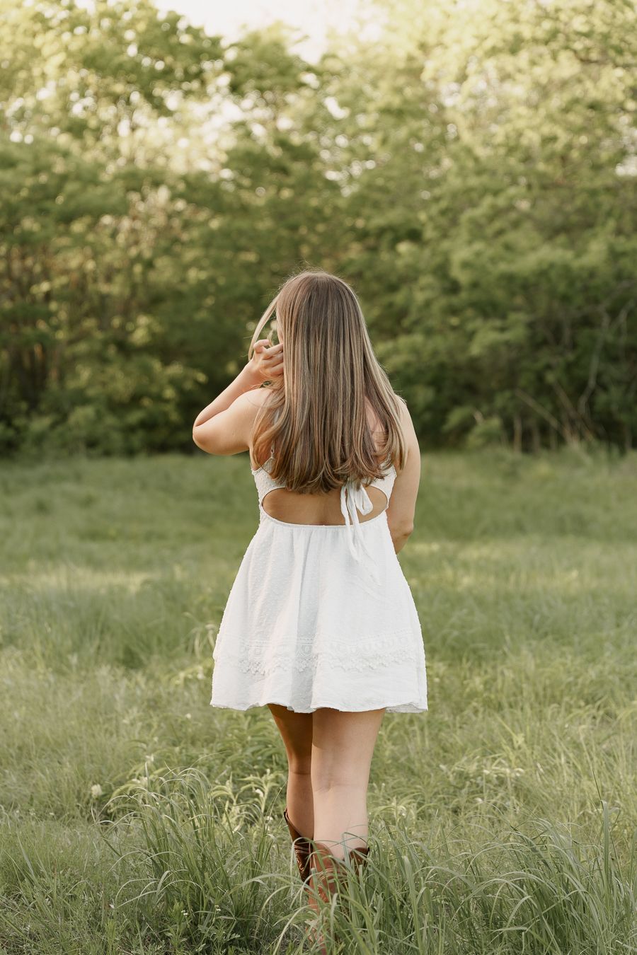 Young woman in white sundress and cowboy boots viewed from behind in lush green field.