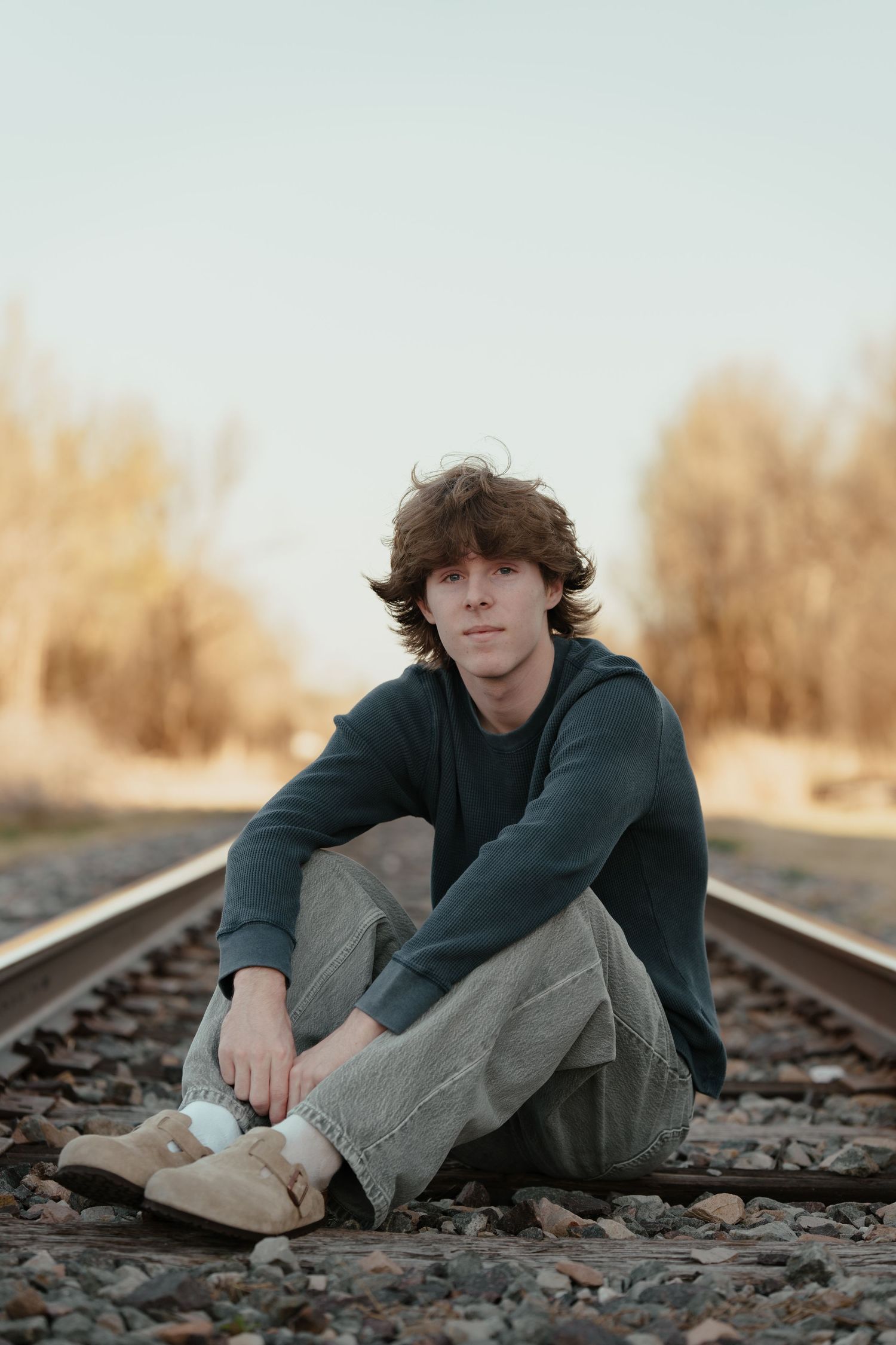 Young man with curly hair sitting on railroad tracks wearing a dark hoodie and jeans in a rural setting.