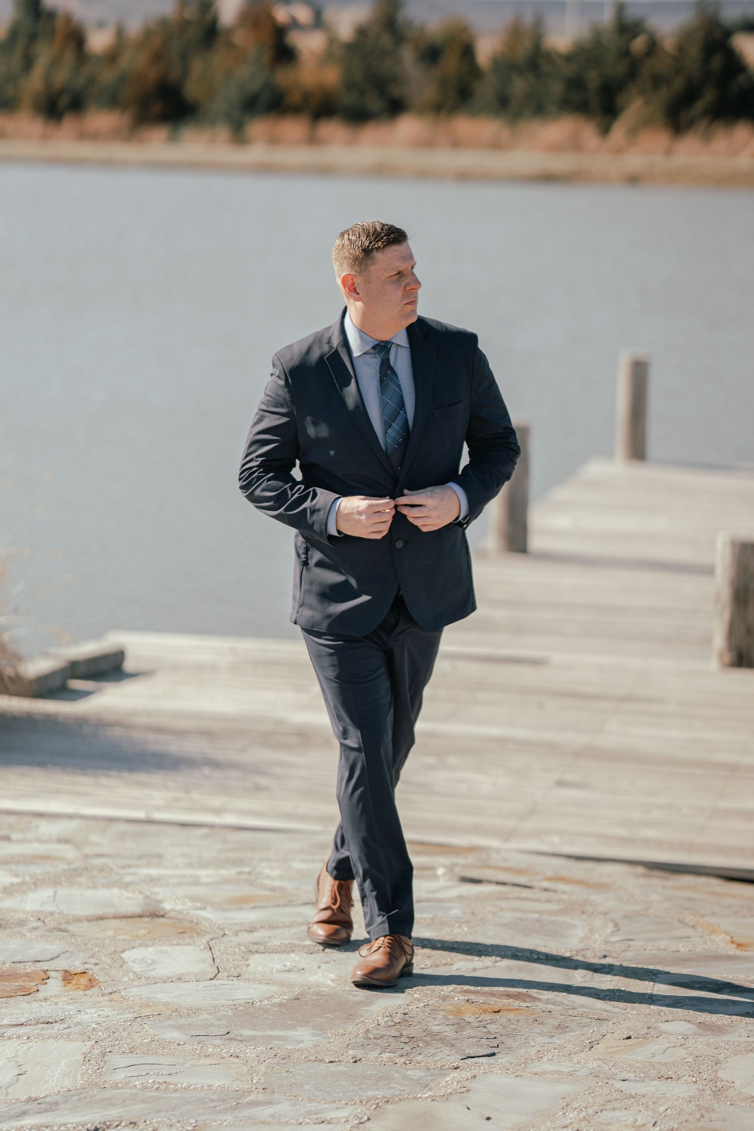 Man in navy suit and tie walking on stone dock by lake, adjusting jacket buttons on sunny day.