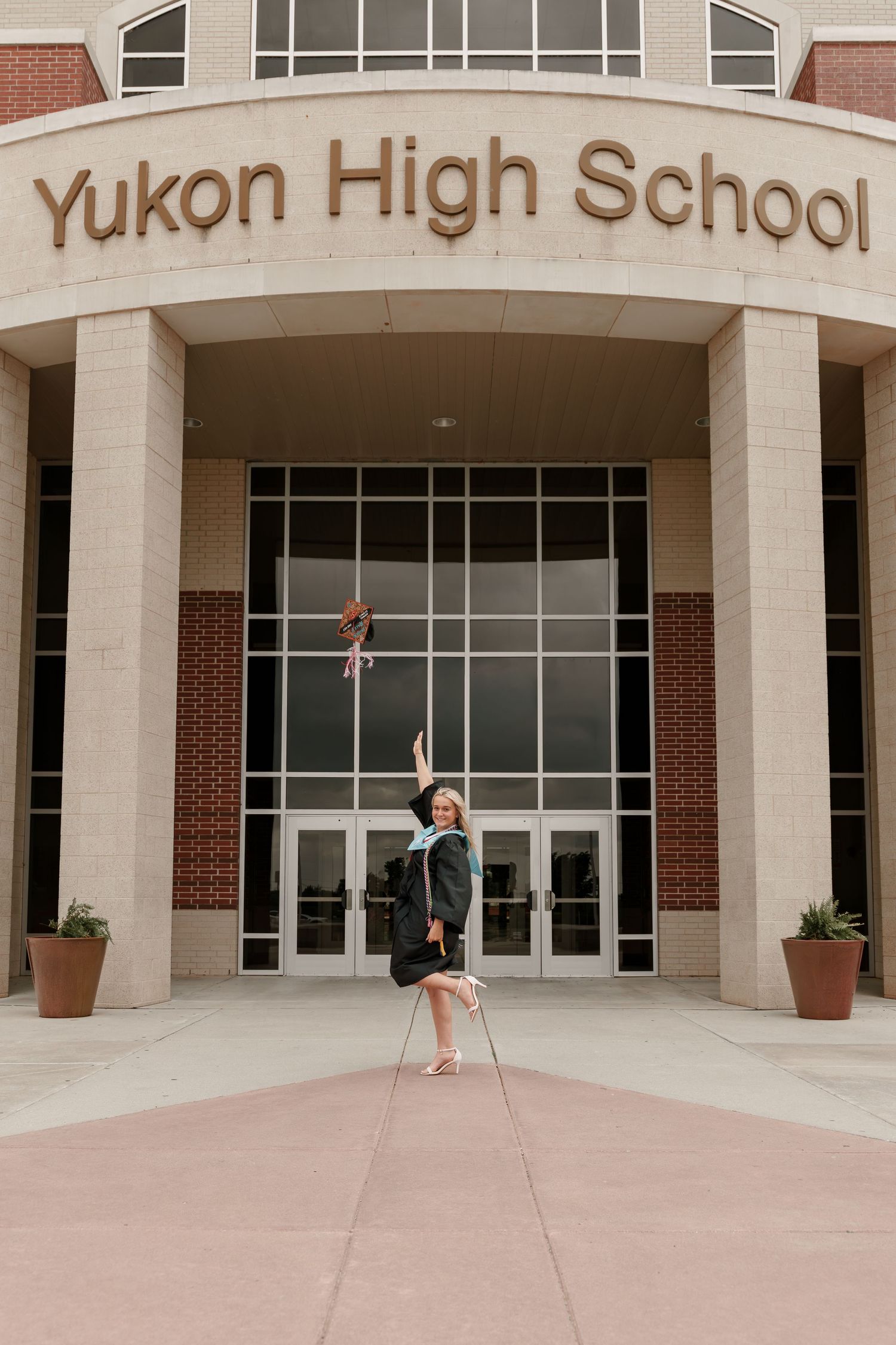 A graduate in cap and gown joyfully jumps outside Yukon High School, tossing their cap in the air.