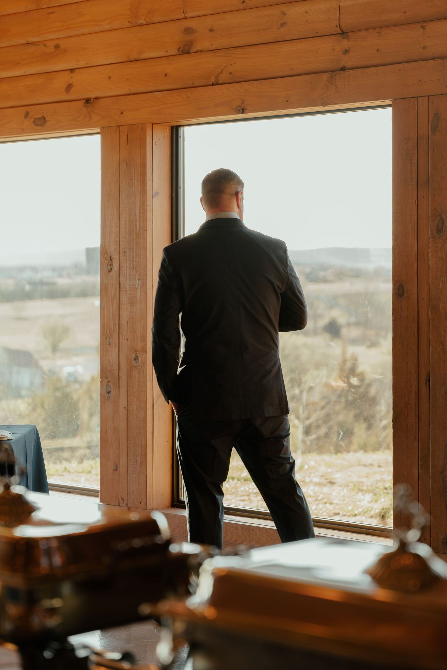 Man in black suit standing at large wooden-framed window overlooking rural countryside landscape in a rustic interior.