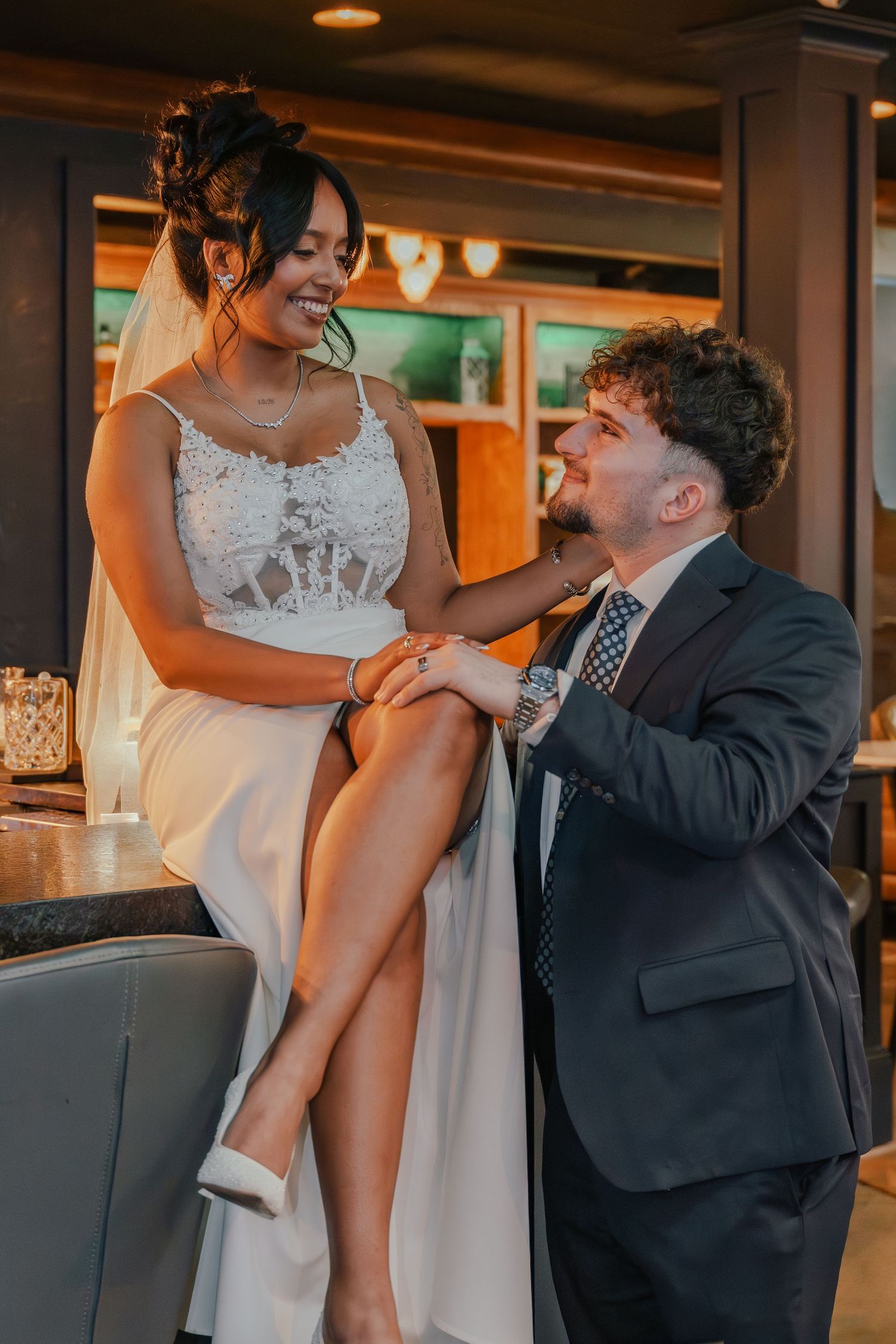 Bride in white lace dress sits on bar stool smiling at groom in navy suit during wedding reception.