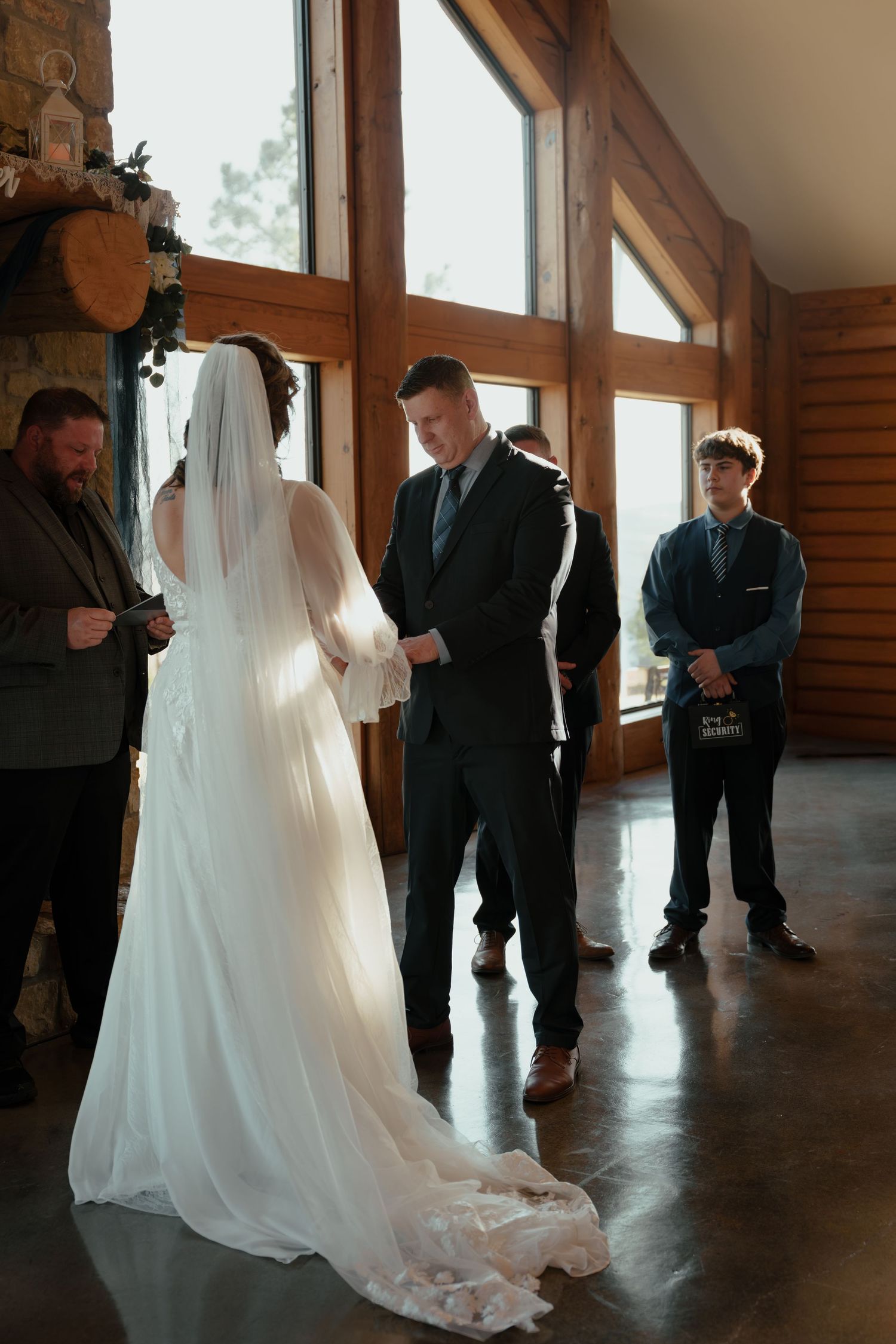 A wedding party stands in a rustic venue with wooden walls and large windows as a bride's white gown trails behind her.