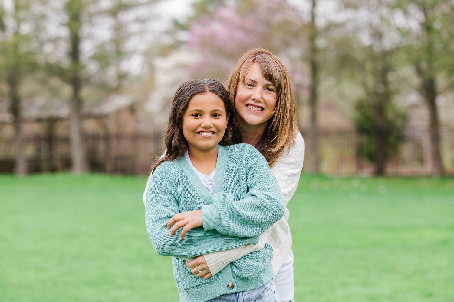 A woman hugs a young girl from behind, both smiling outdoors on green grass with blooming trees in spring.