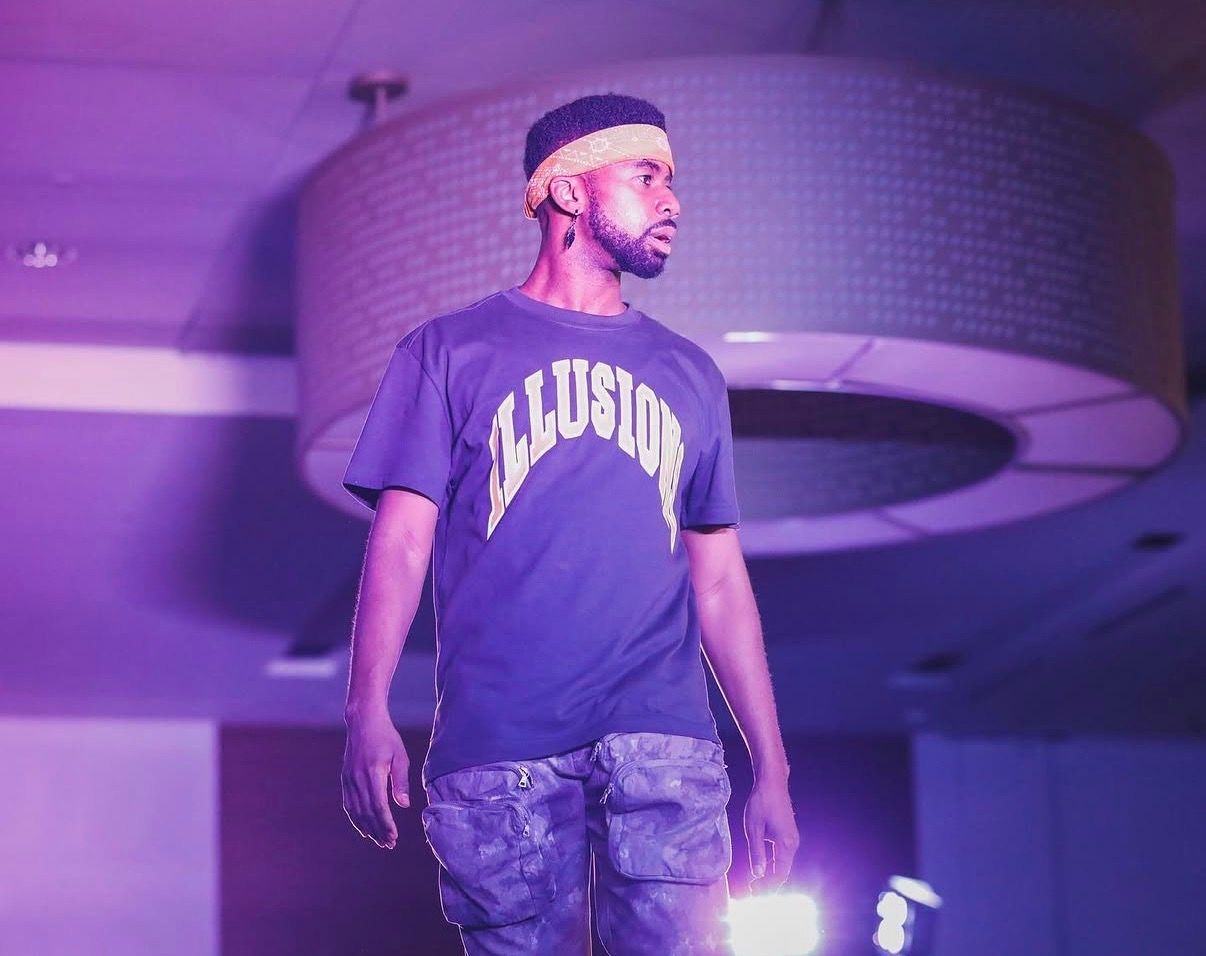 A performer in a dark t-shirt and camo shorts stands on stage under purple lighting during a live show.
