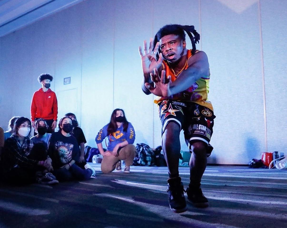 Dancer performs energetic moves in front of seated audience during indoor dance performance.