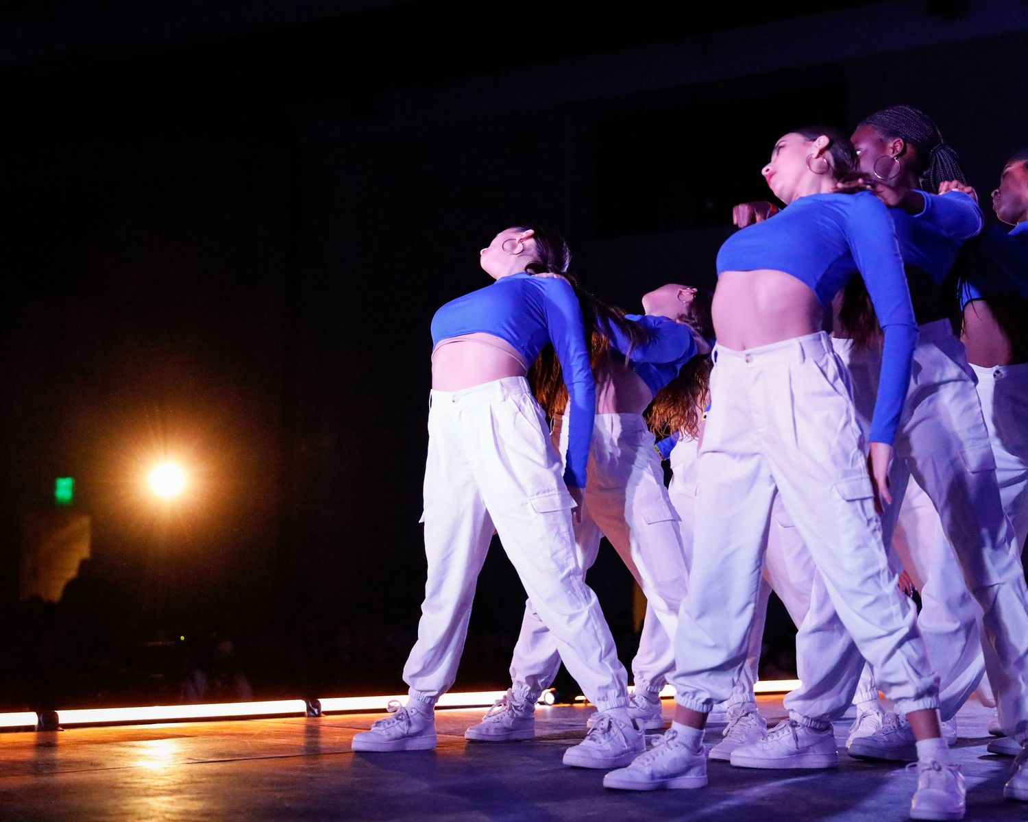 Dancers in white outfits perform a synchronized dance routine on a dimly lit stage during an evening performance.