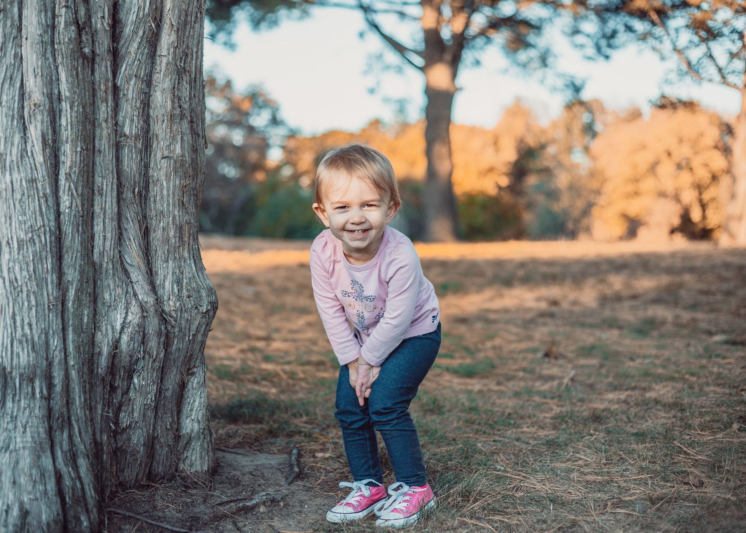A person in a purple sweater crouches and smiles in a forest setting during golden hour sunset.