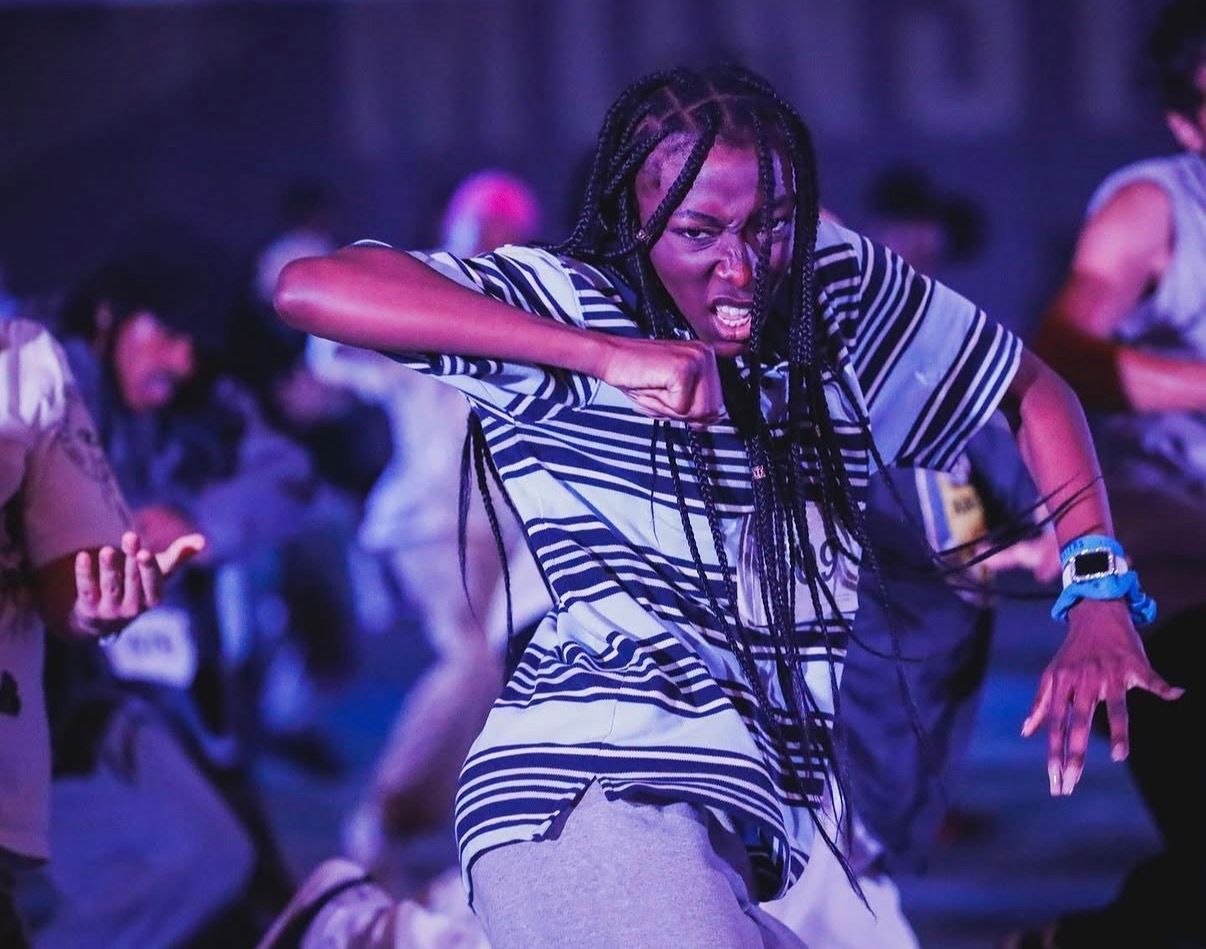 Performers in striped shirts dance energetically on stage under purple and blue lighting during a live show.