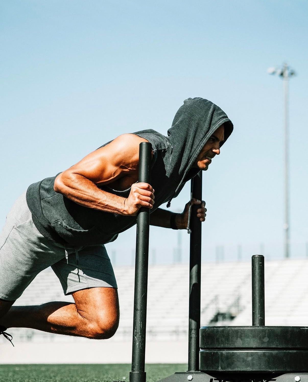 Athletic figure in dark hoodie pushing weighted sled during intense outdoor workout on turf field.