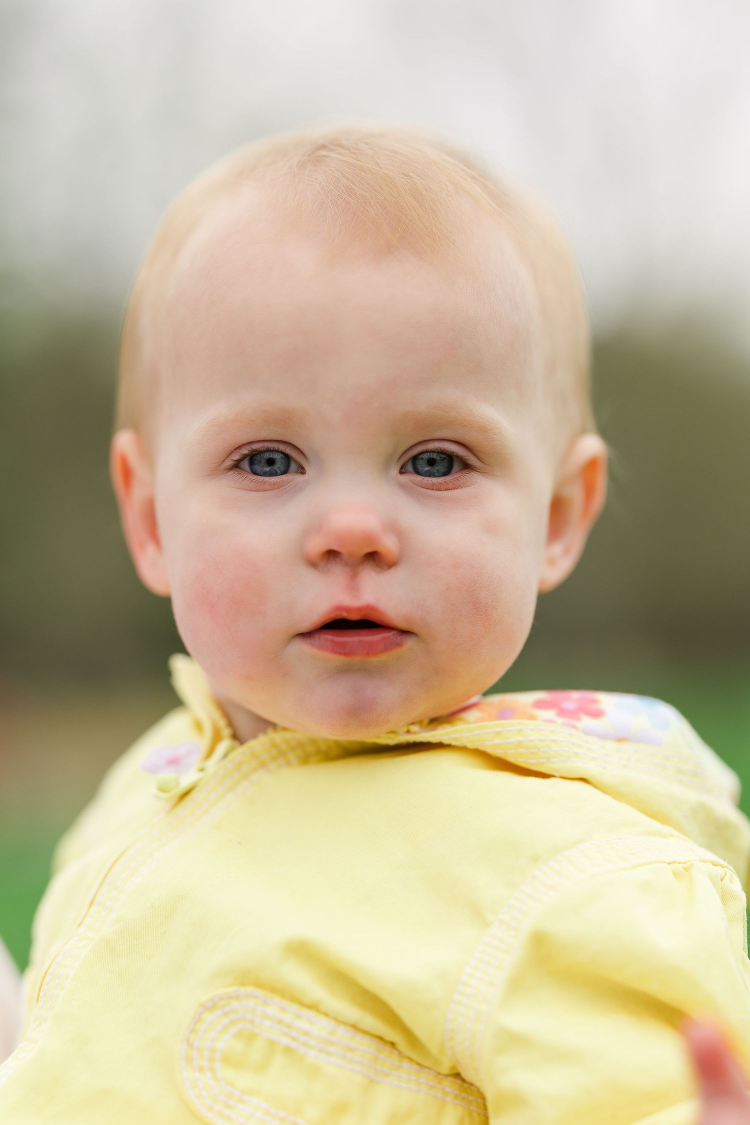 Adorable baby in yellow floral jacket with blue eyes and rosy cheeks outdoors in natural light.