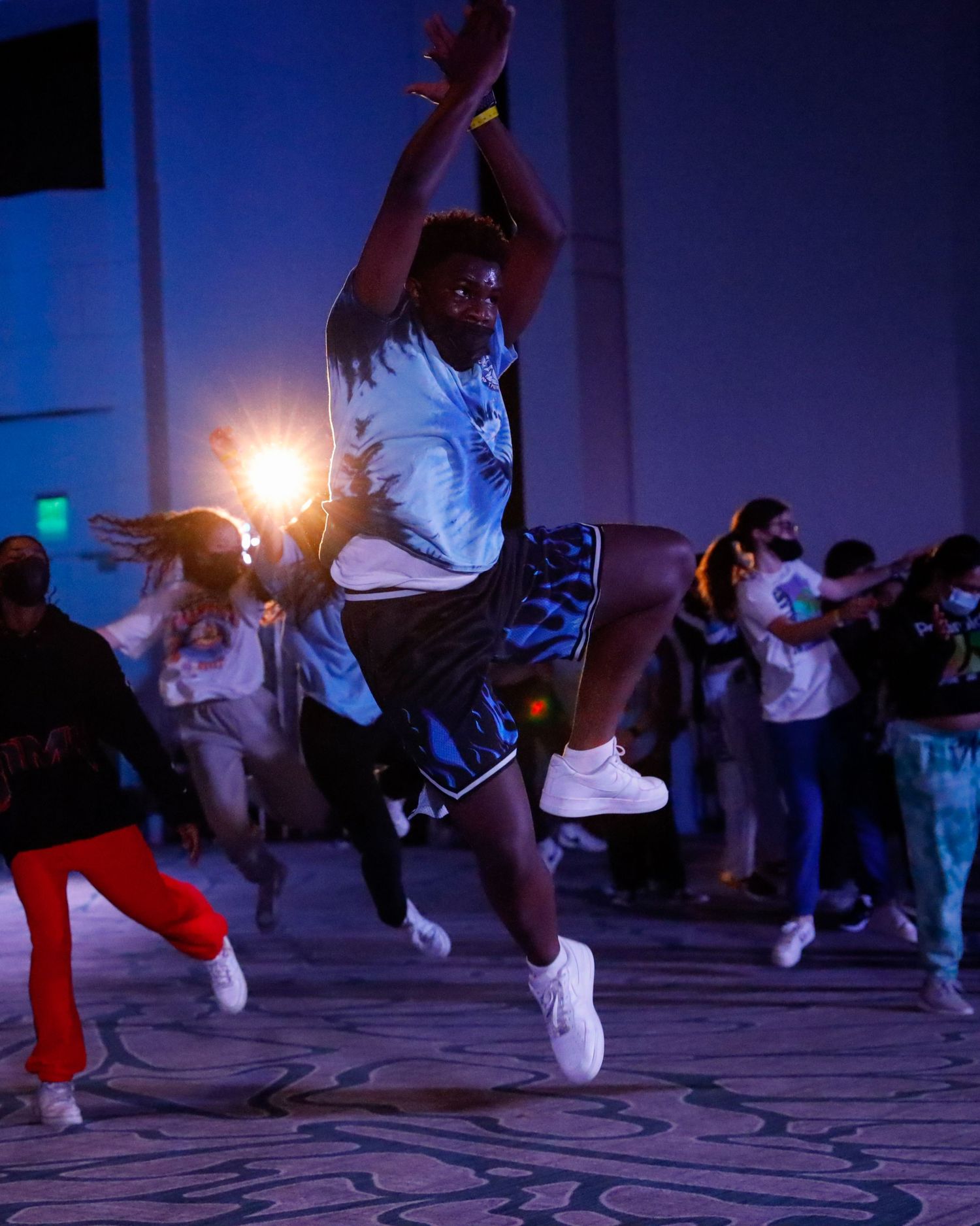 Dancers perform energetic hip hop moves in a dimly lit studio with blue lighting and glowing effects.