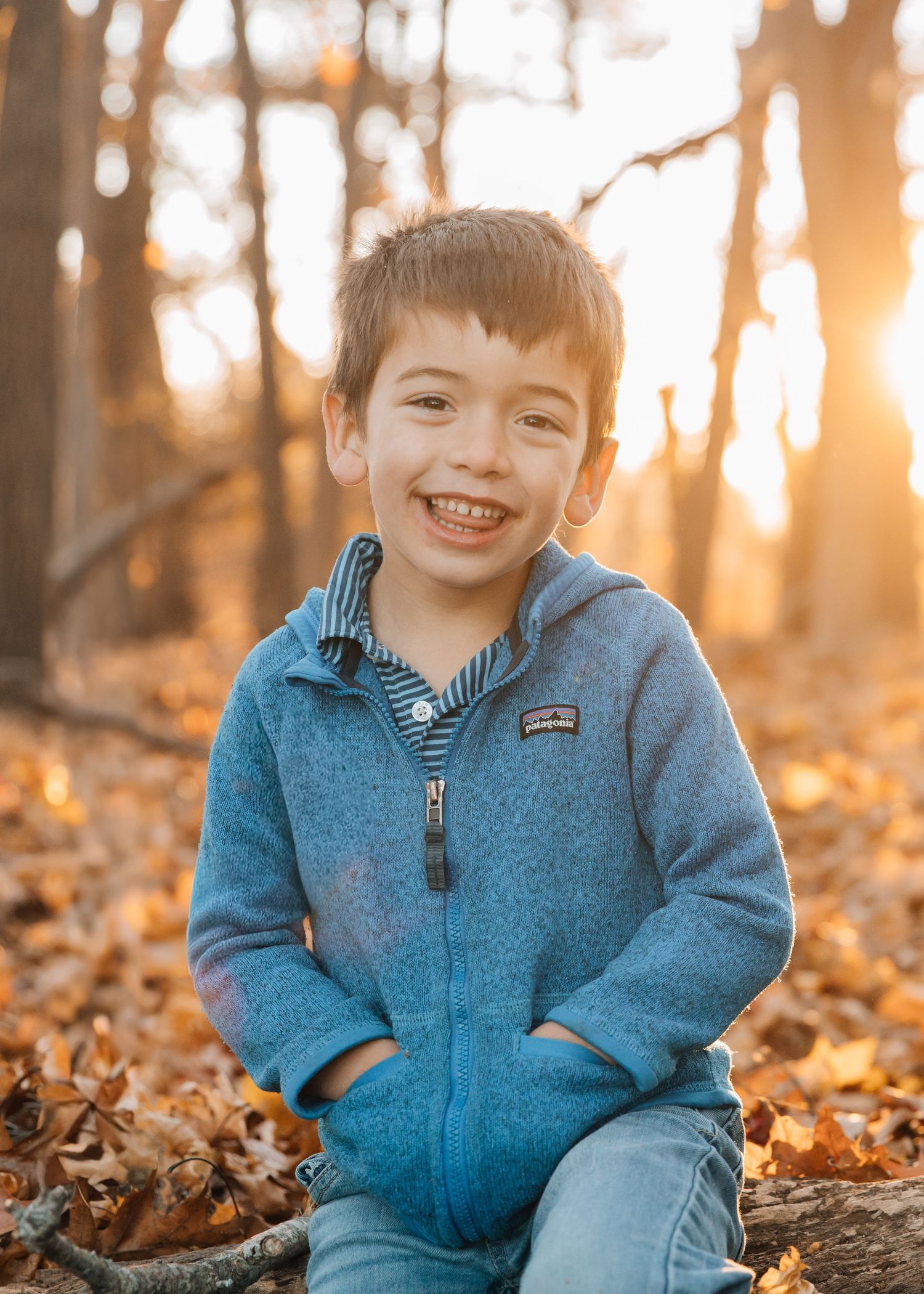 Child wearing blue fleece jacket plays in fallen autumn leaves at sunset in a wooded area.