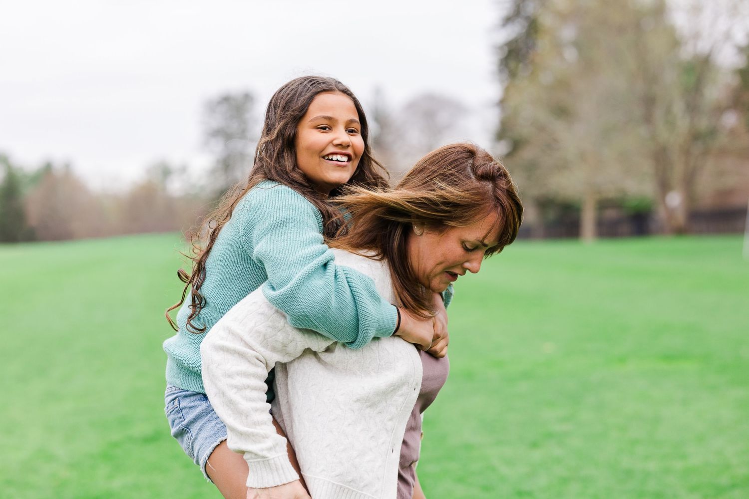 A young girl laughs while getting a piggyback ride from a woman in a green park setting.