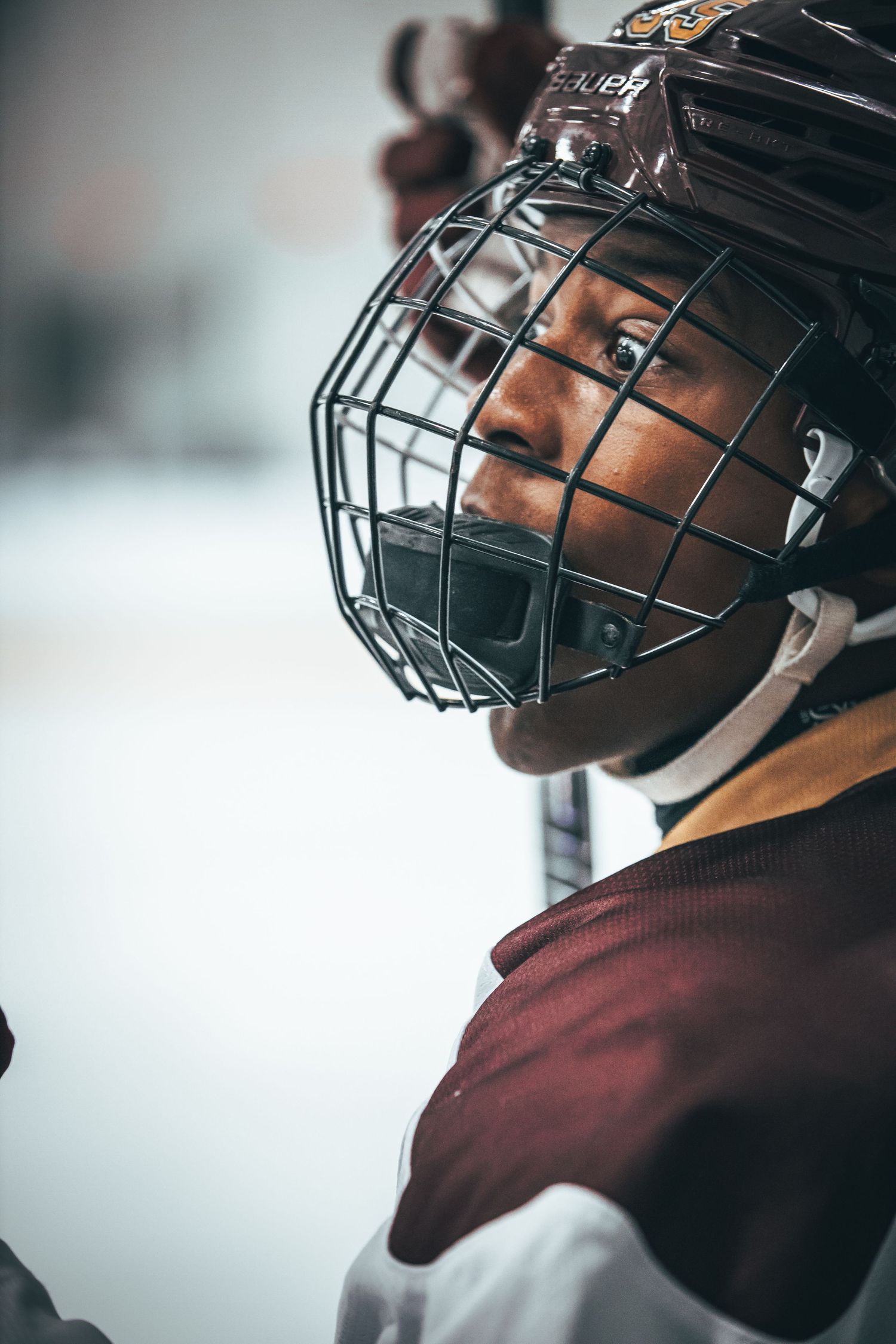 Hockey player wearing maroon jersey and protective helmet with cage viewed in profile against blurred background.