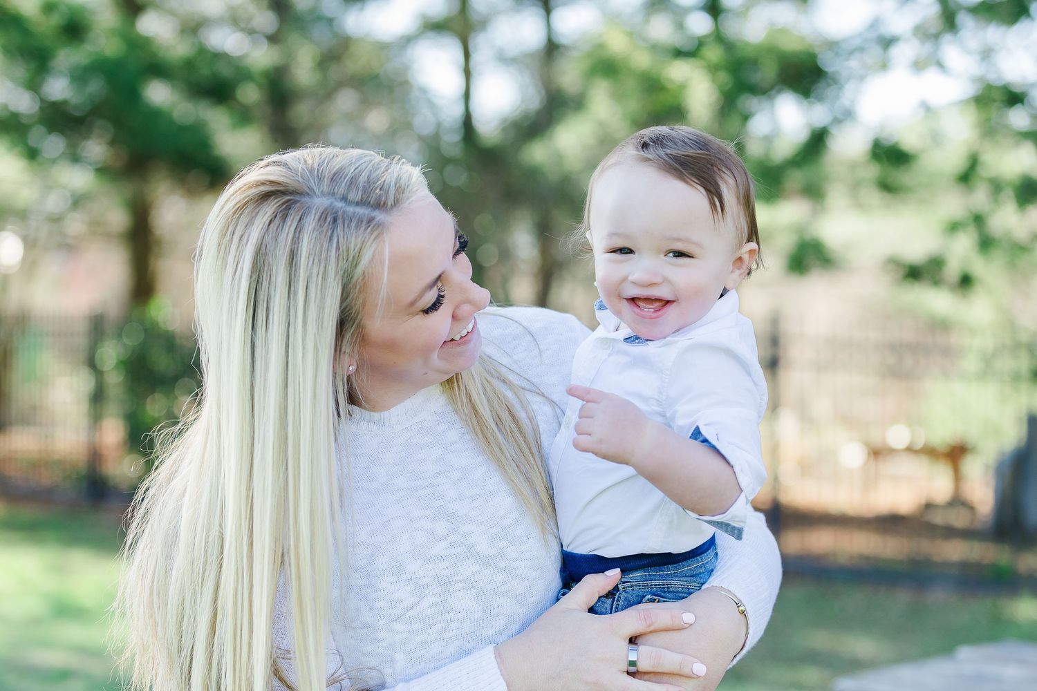 A smiling blonde woman holds a happy laughing baby boy outdoors in a green park setting.