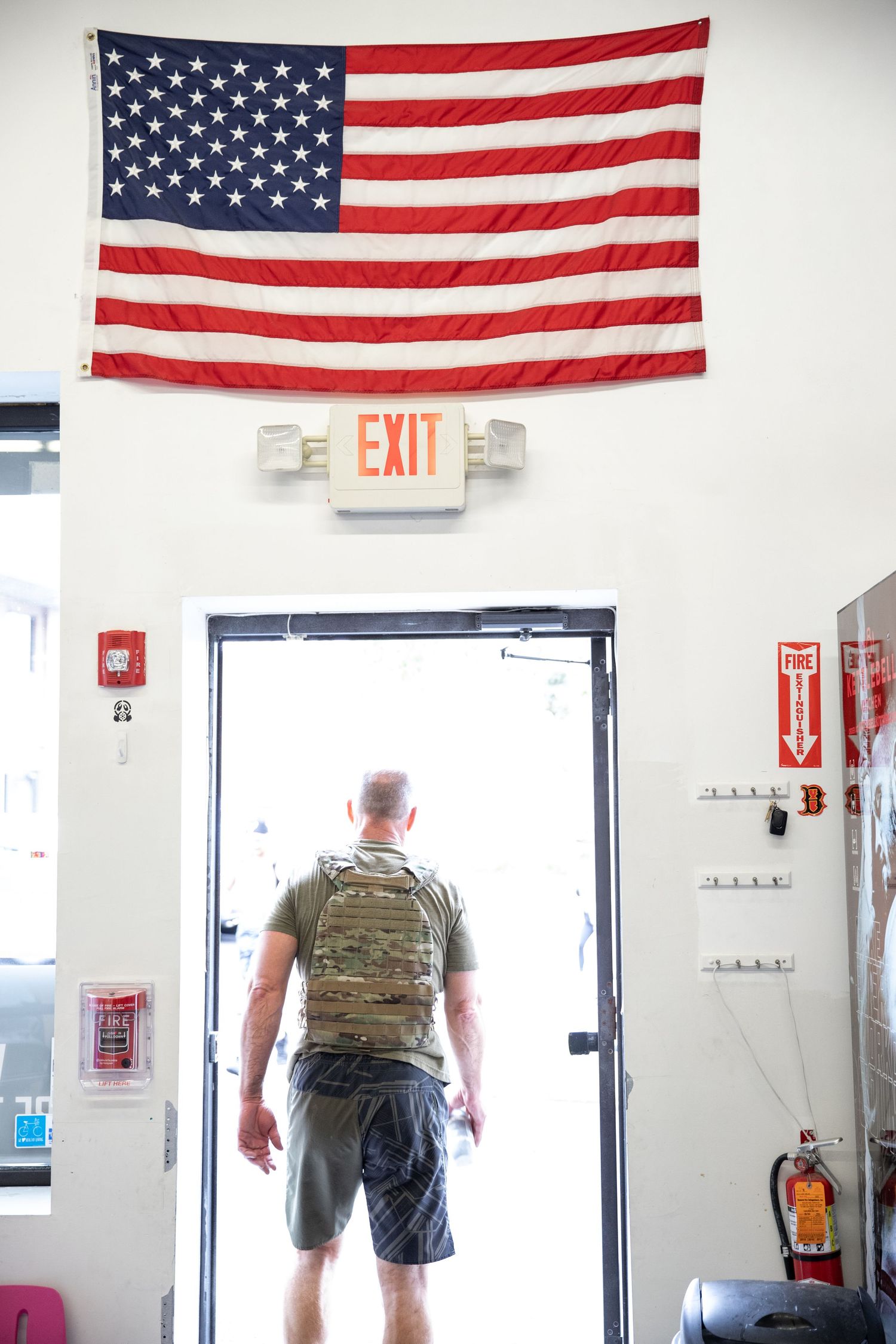 American flags hang above doorways in a white hallway with a visible exit sign and tactical gear-wearing personnel.