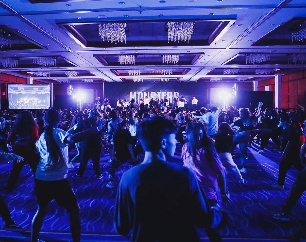 Large crowd of people dancing at a nightclub event with blue lighting and chandeliers overhead.