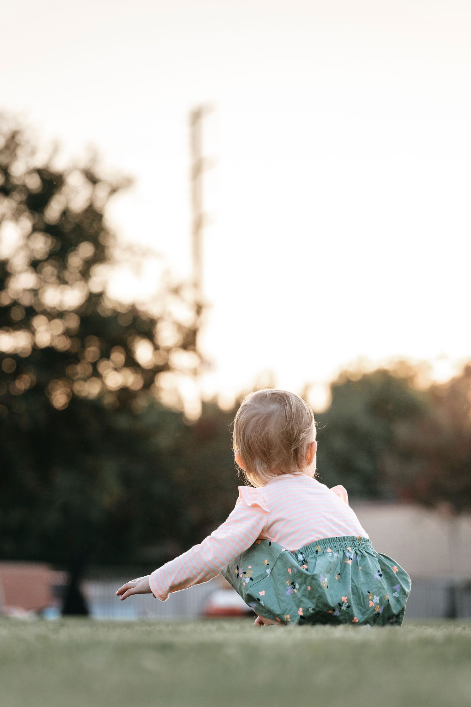 Small child in teal dress crawling on grassy field during golden hour.