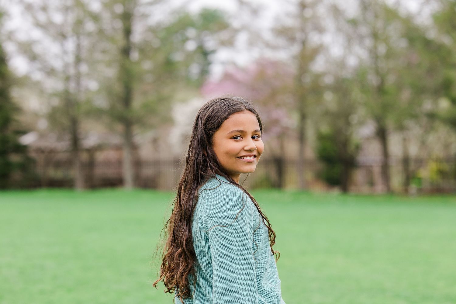 A smiling young girl with long dark hair wearing a light blue jacket stands in a lush green park with trees behind her.