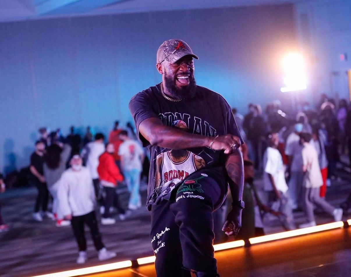 Performer dances enthusiastically under stage lighting at an indoor event with crowd in background.