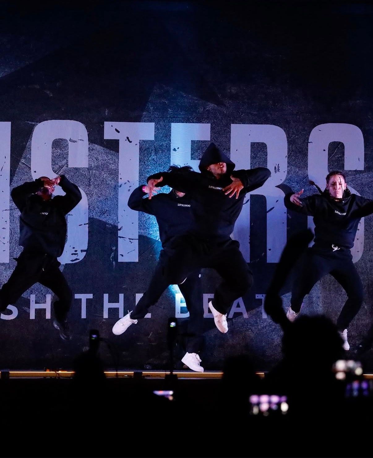 Silhouetted dancers perform synchronized movements against a dark backdrop with illuminated text behind them.