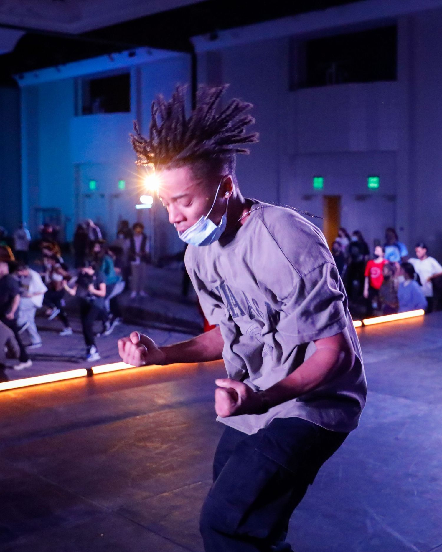 A dancer in grey performs a dynamic movement sequence under dramatic blue lighting on a dark stage.