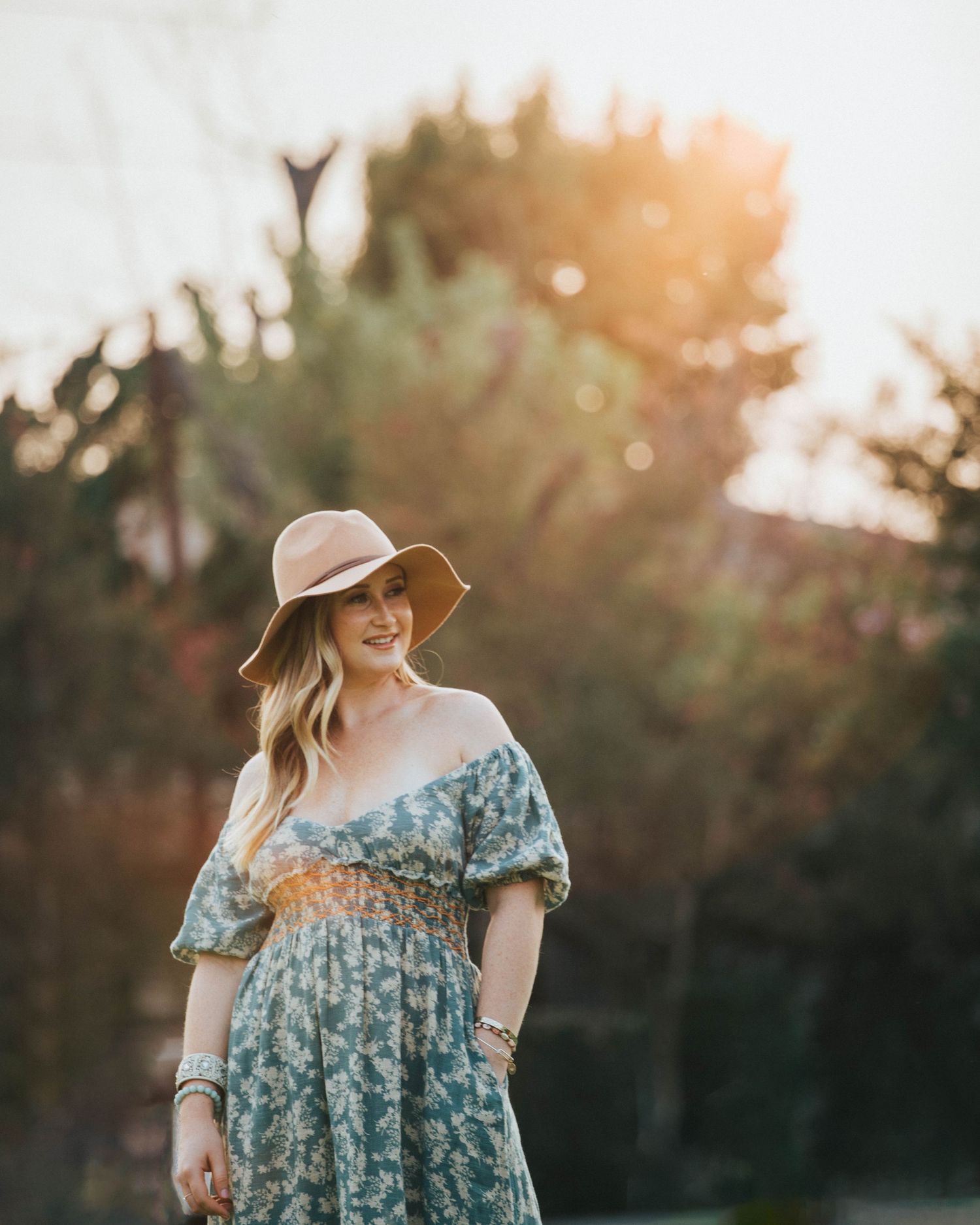 Woman in floral dress and sun hat poses in garden during golden hour sunset.