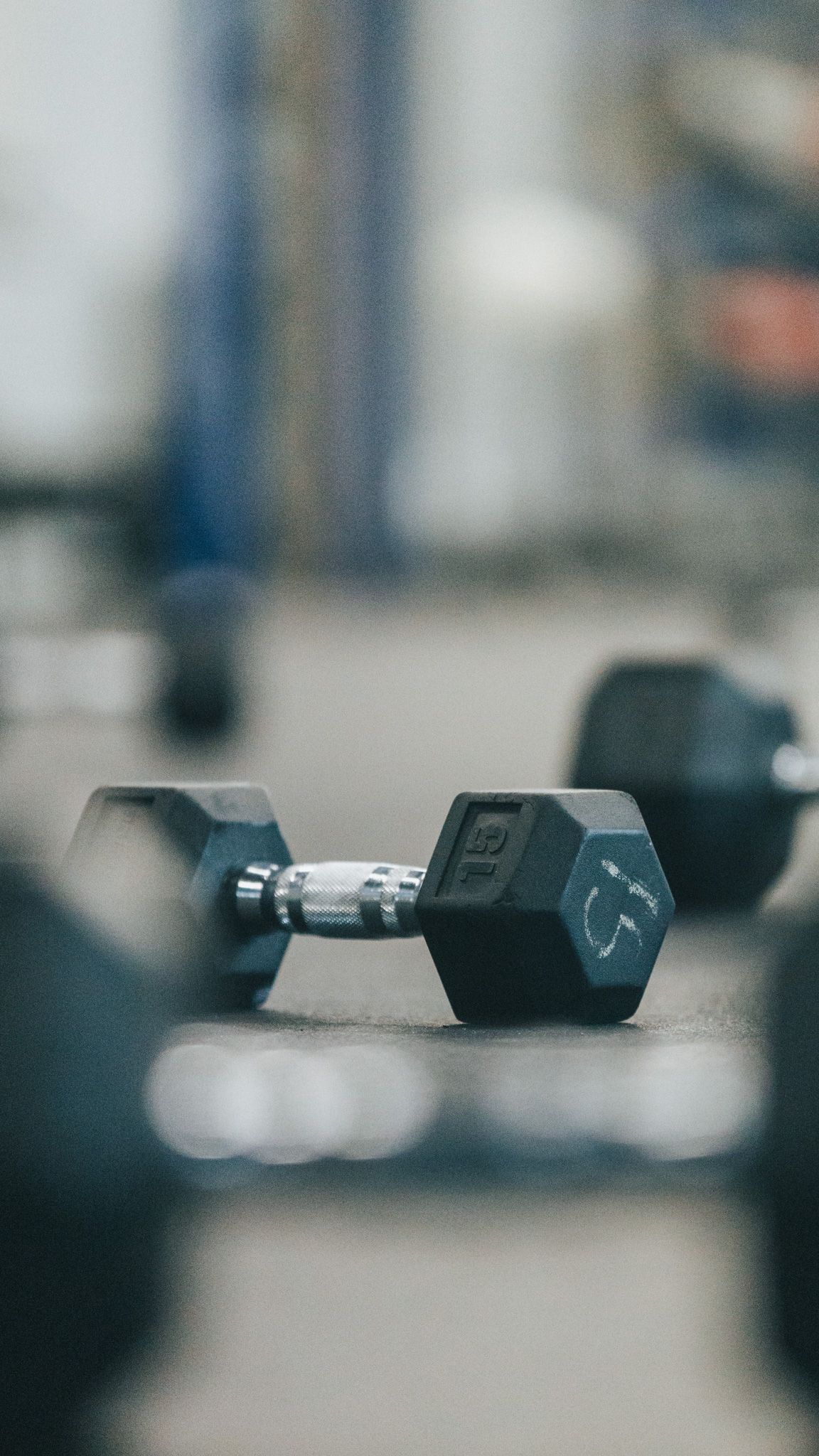 Black hexagonal dumbbell resting on a blurred gym floor with soft bokeh effect in the background.