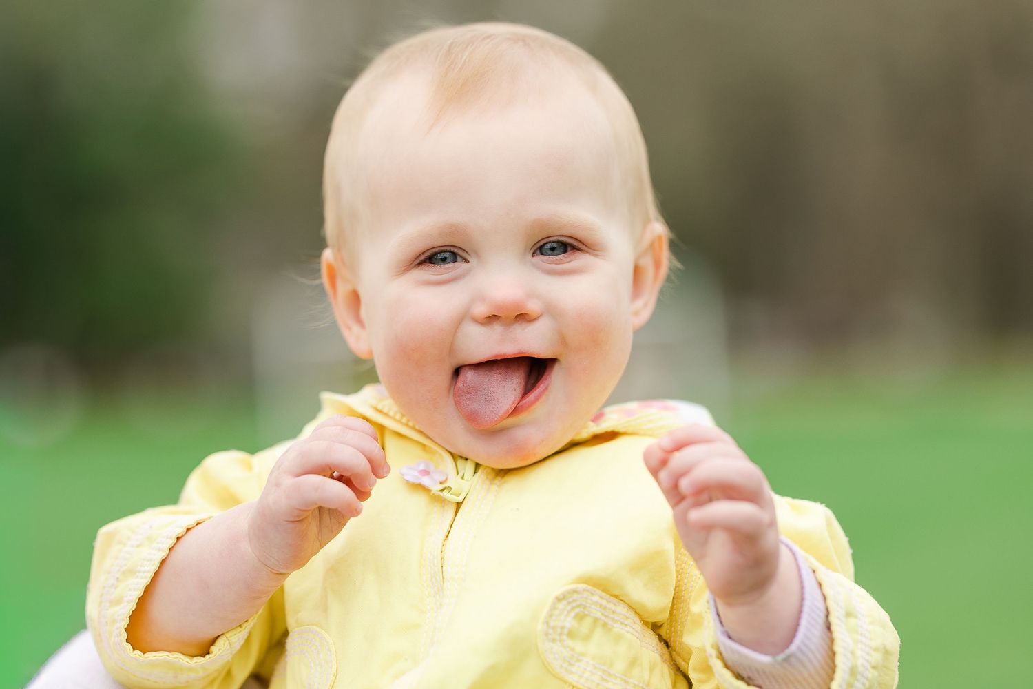 Adorable baby in yellow outfit smiling with tongue out outdoors.