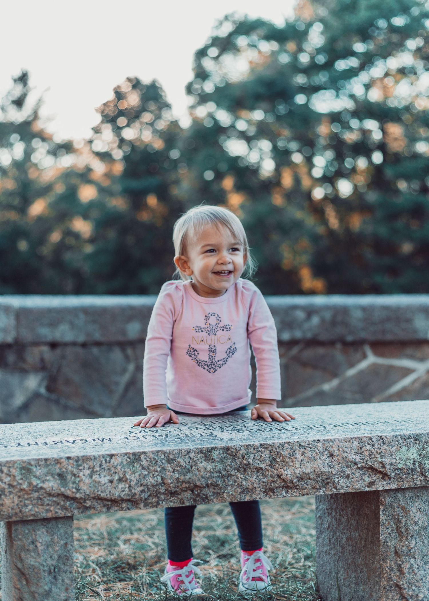 Child in pink sweater sits playfully on concrete ledge during golden hour in outdoor setting.