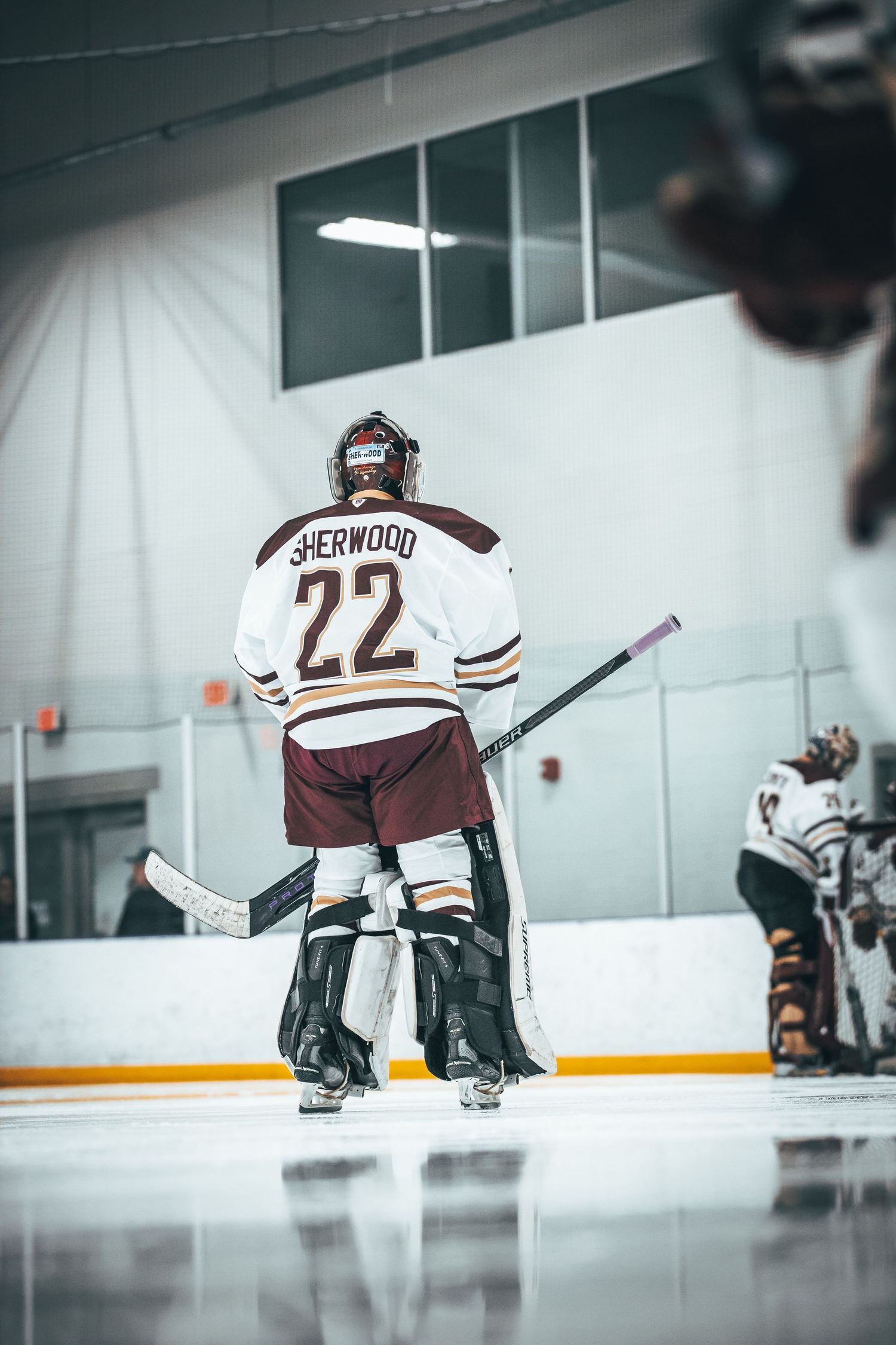 Hockey goalie in brown and white jersey crouches in defensive position on ice rink during practice session.