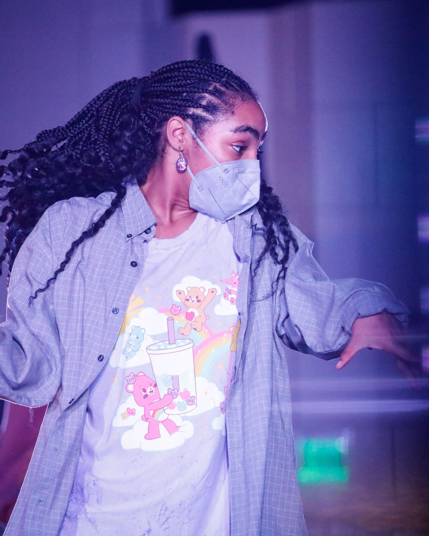 A performer in a gray shirt with long braids dances in dramatic purple lighting on stage.