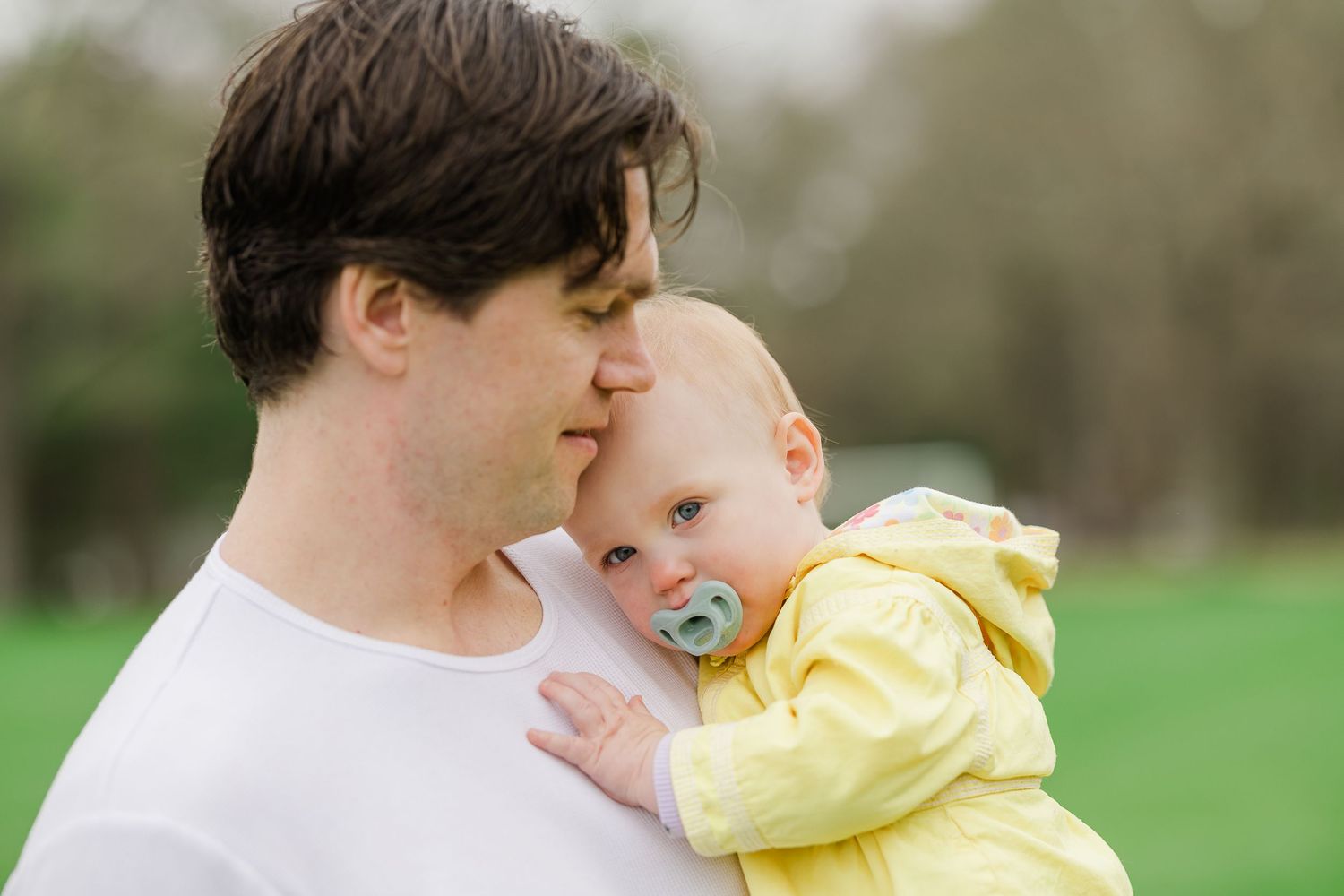 A father tenderly kisses his baby in a yellow hoodie with a pacifier outdoors in a green park.
