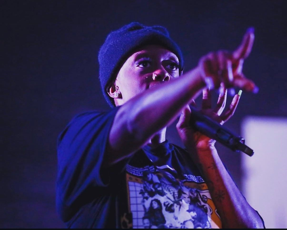 A performer in a blue beanie gestures energetically while holding a microphone during a concert performance.