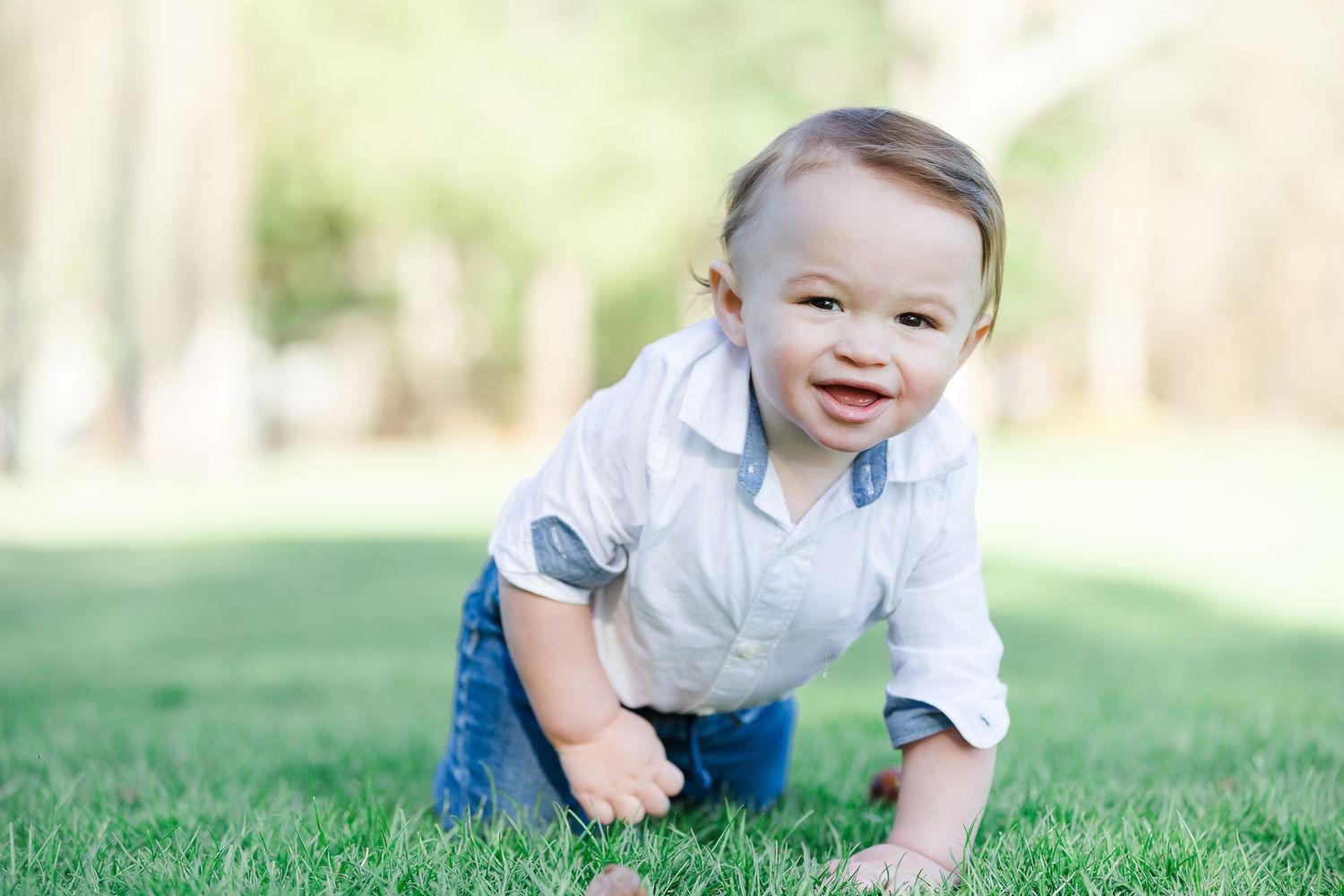 A cheerful baby boy in a white shirt and jeans crawling on green grass outdoors in a sunny park.