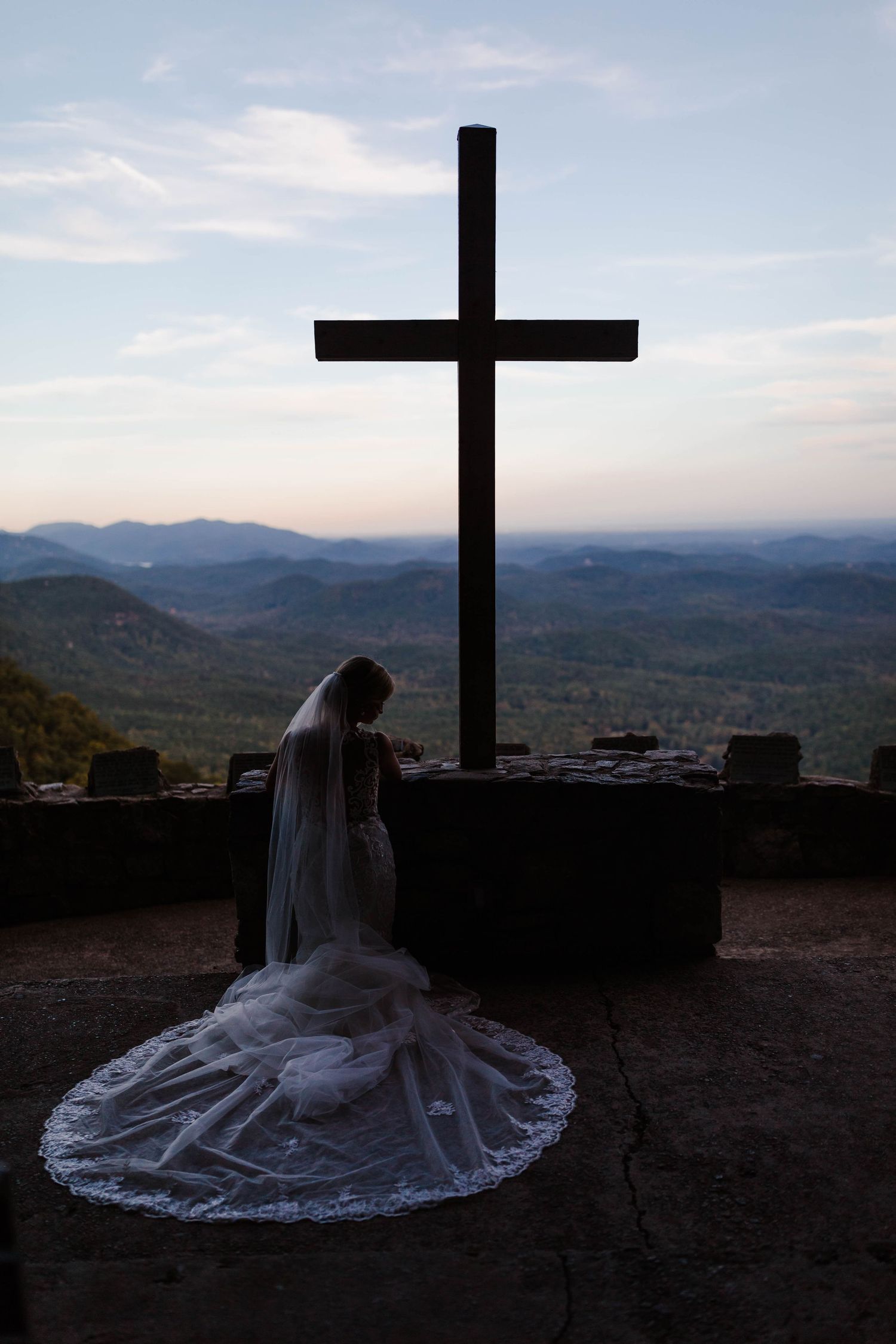 A figure in a white dress kneels before a large cross at sunset overlooking a scenic mountain landscape.