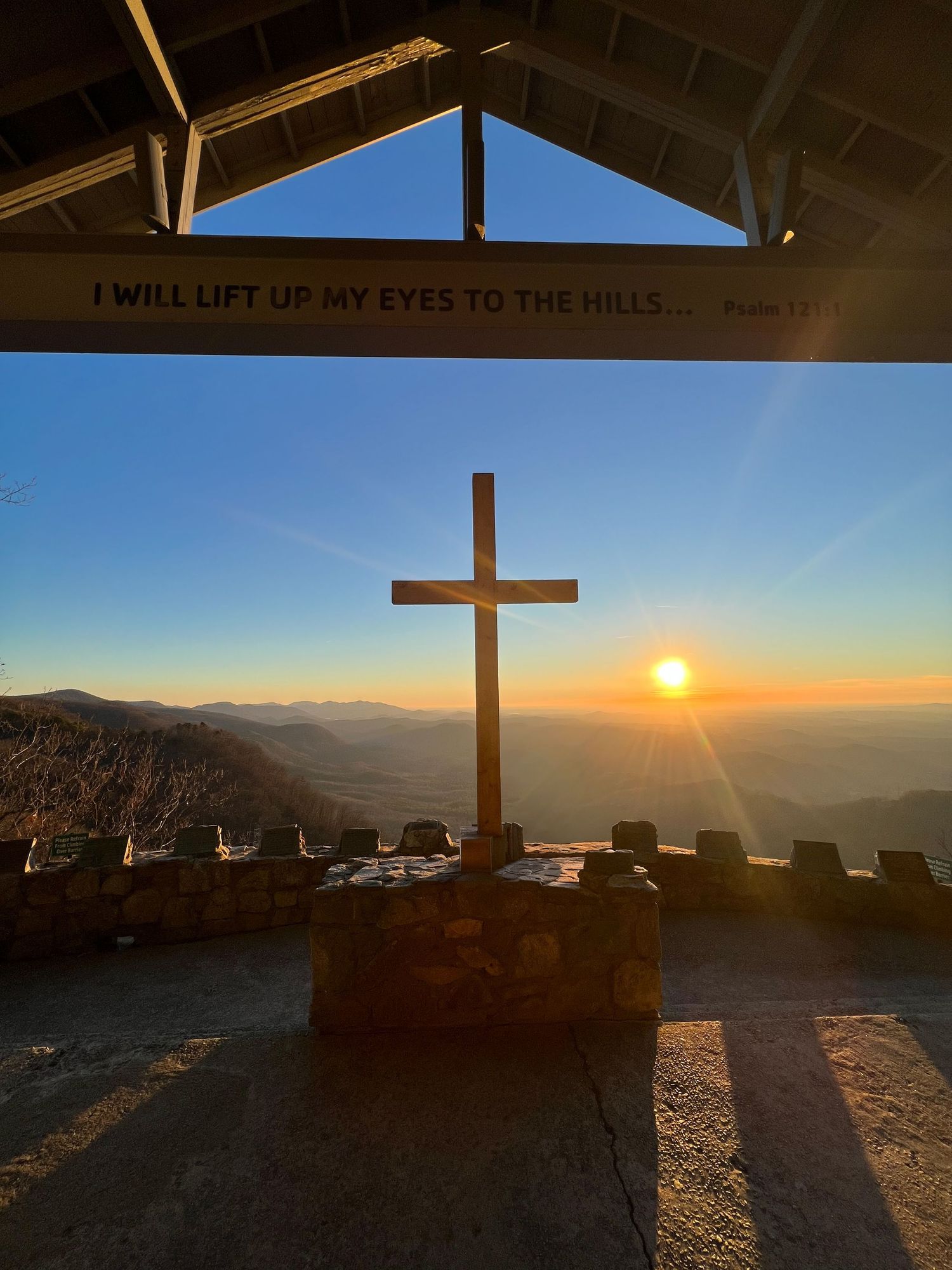 A wooden cross silhouetted against a vibrant sunset over mountain peaks, with 'I will lift up my eyes to the hills' text above.