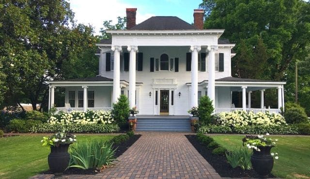 A classic white antebellum style mansion with columns and wraparound porch sits surrounded by lush green landscaping.