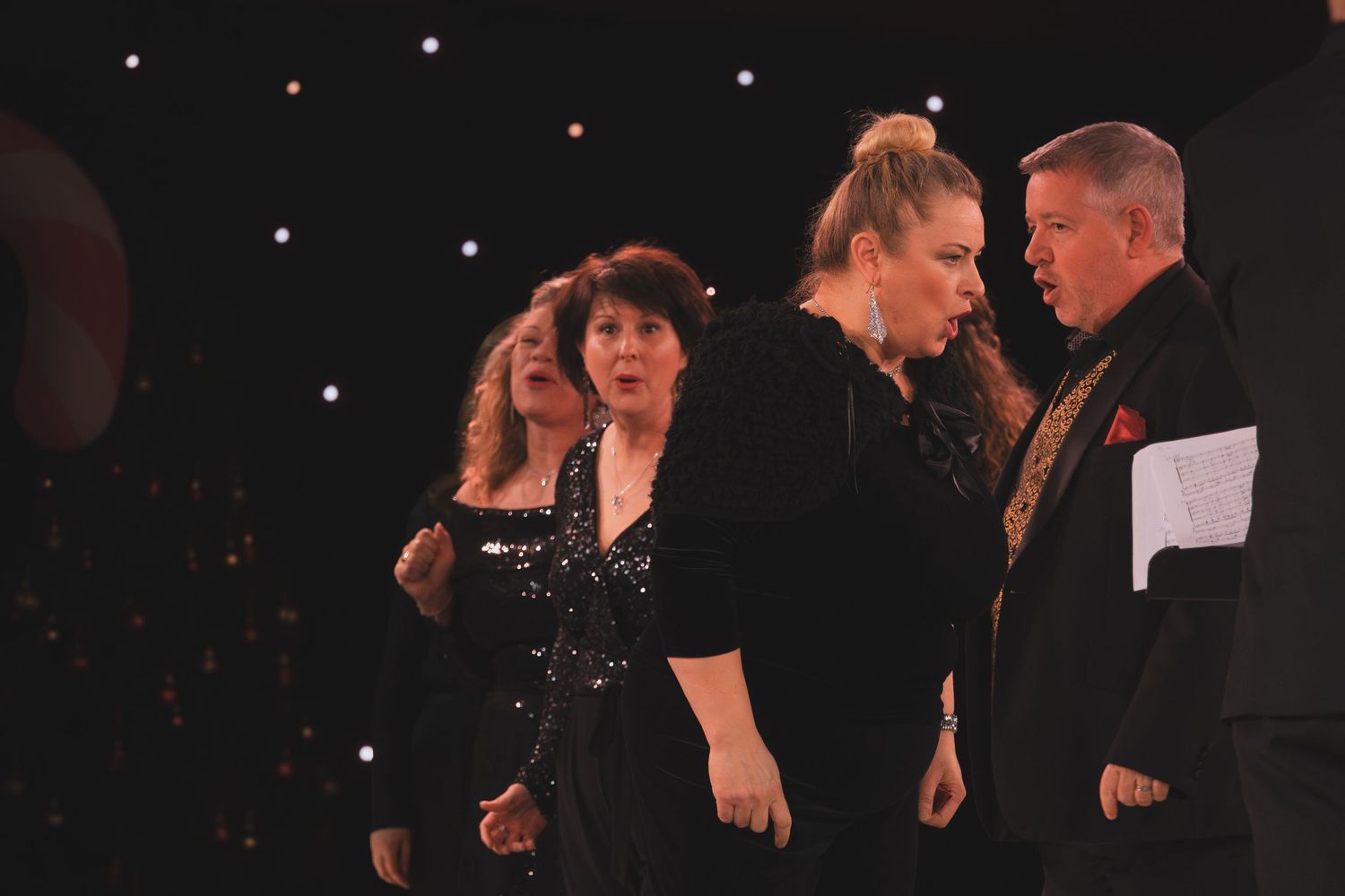 Stage performers in black clothing stand together under dramatic lighting.